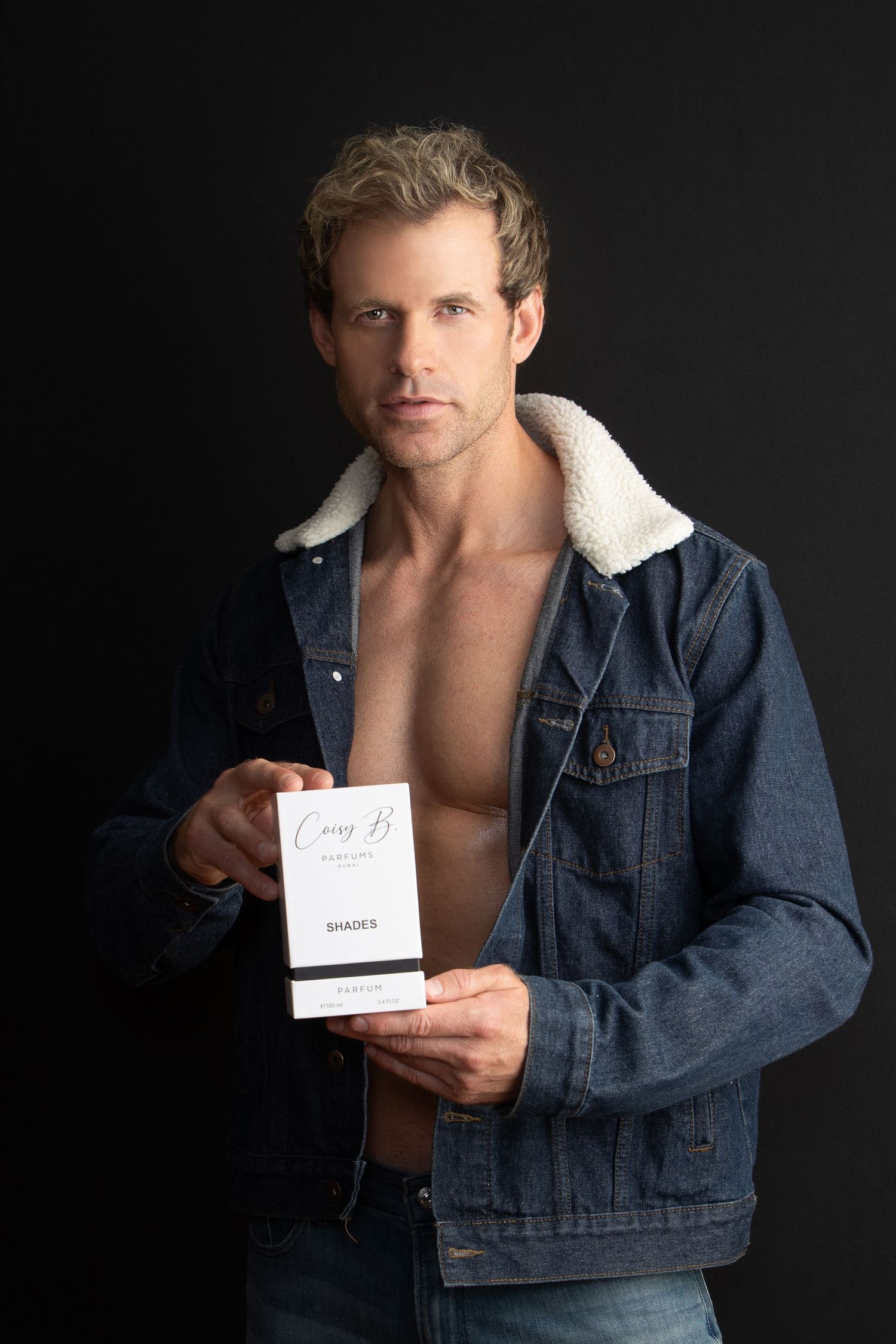 A figure in a denim jacket with sherpa lining poses against a dark background while holding a small white card.