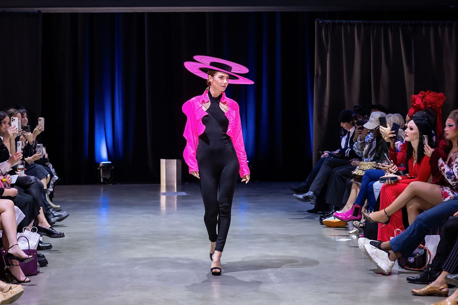 A model walks the runway wearing a dramatic pink sculptural hat and black outfit during a fashion show.