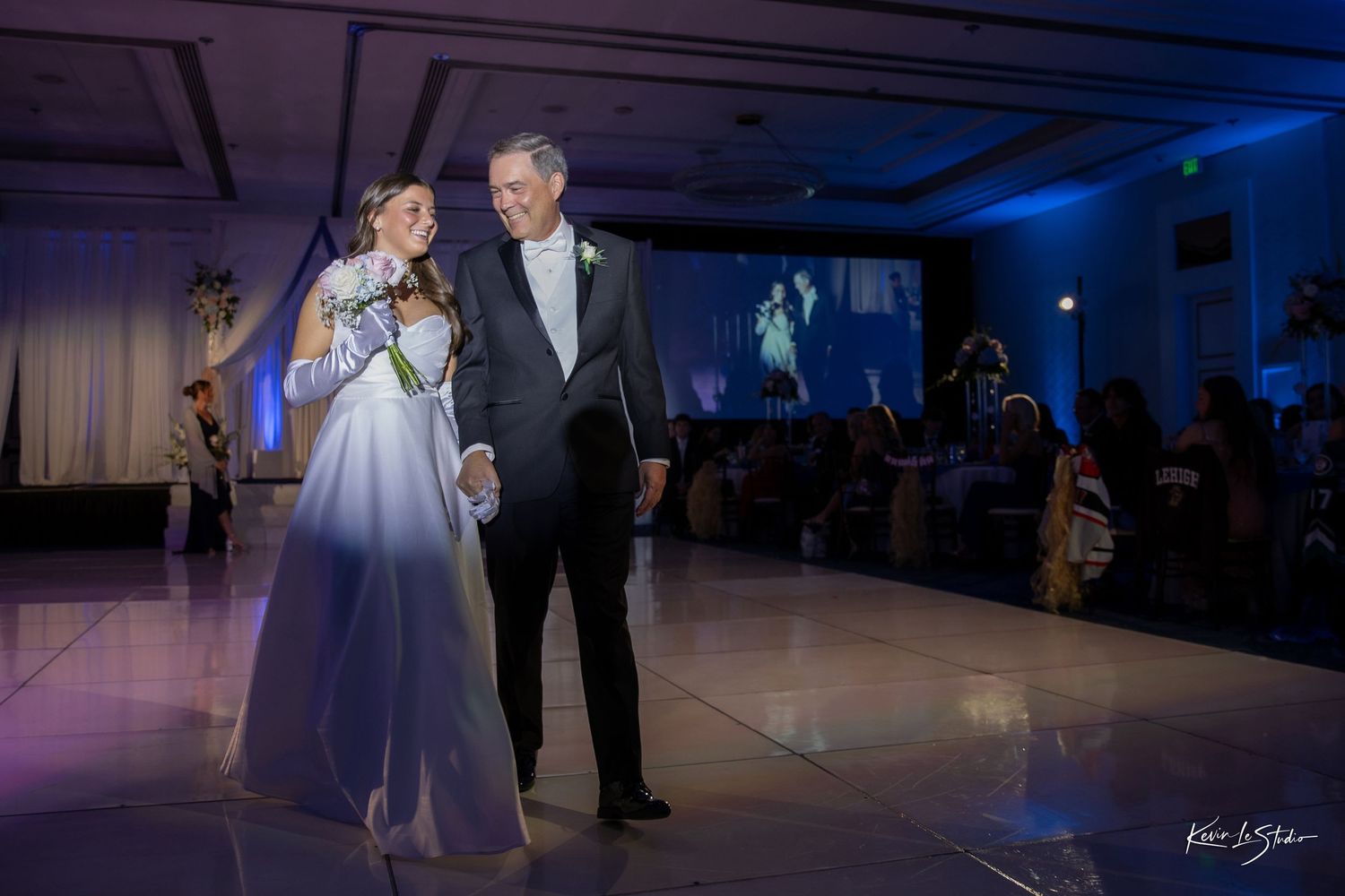 A couple shares a romantic first dance in a ballroom illuminated by blue lighting and surrounded by guests.