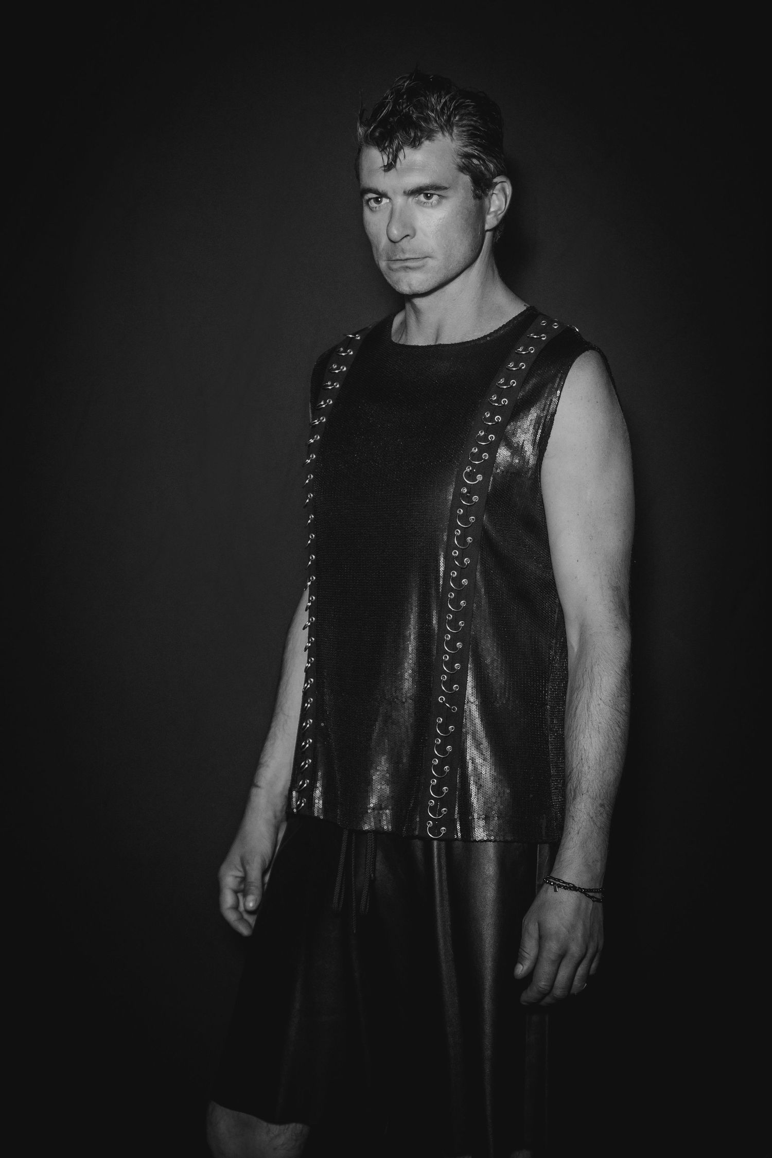 Black and white portrait of person in sleeveless leather top with studded details against dark background.