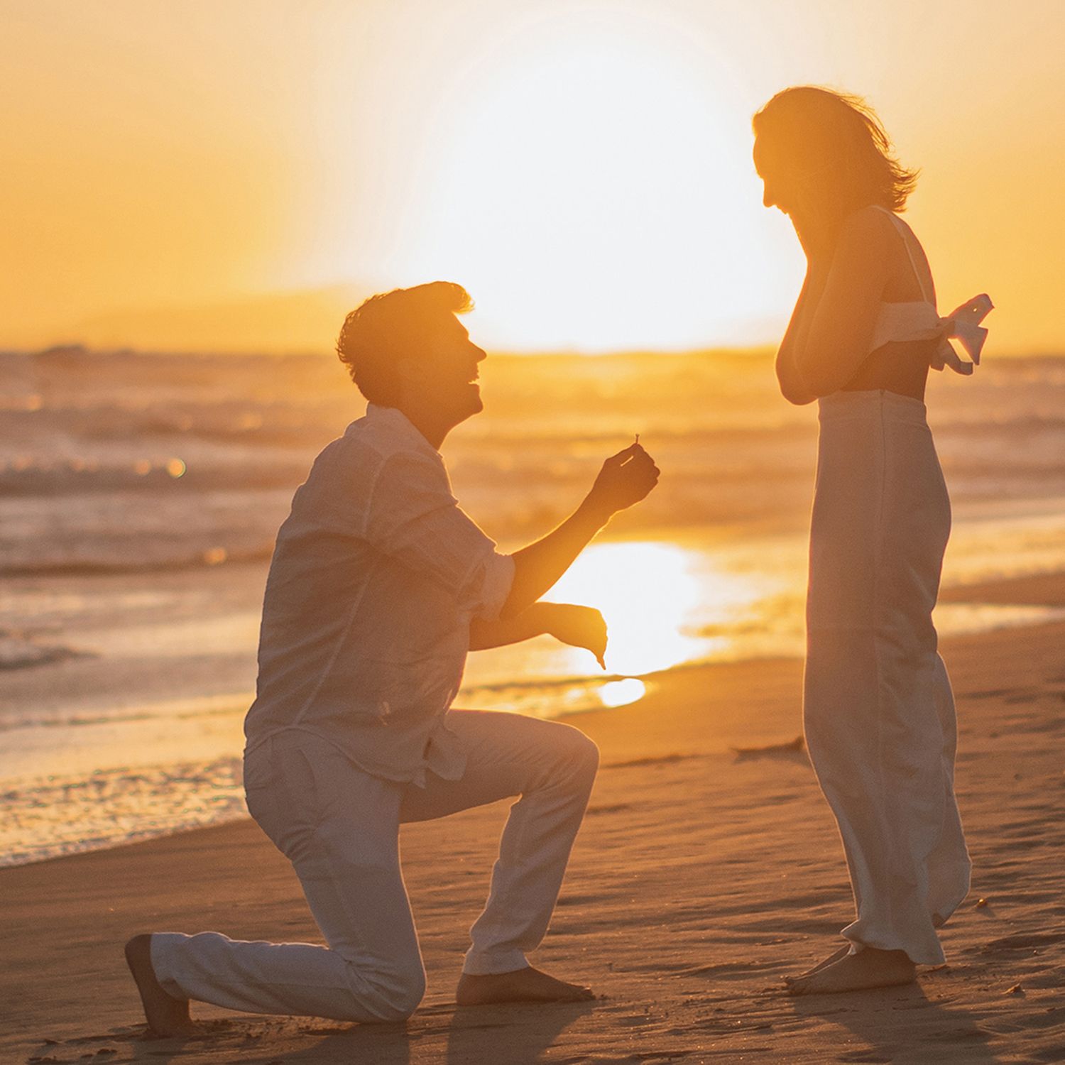 Man proposes to woman on beach at sunset, kneeling with ring while waves reflect golden light behind them.