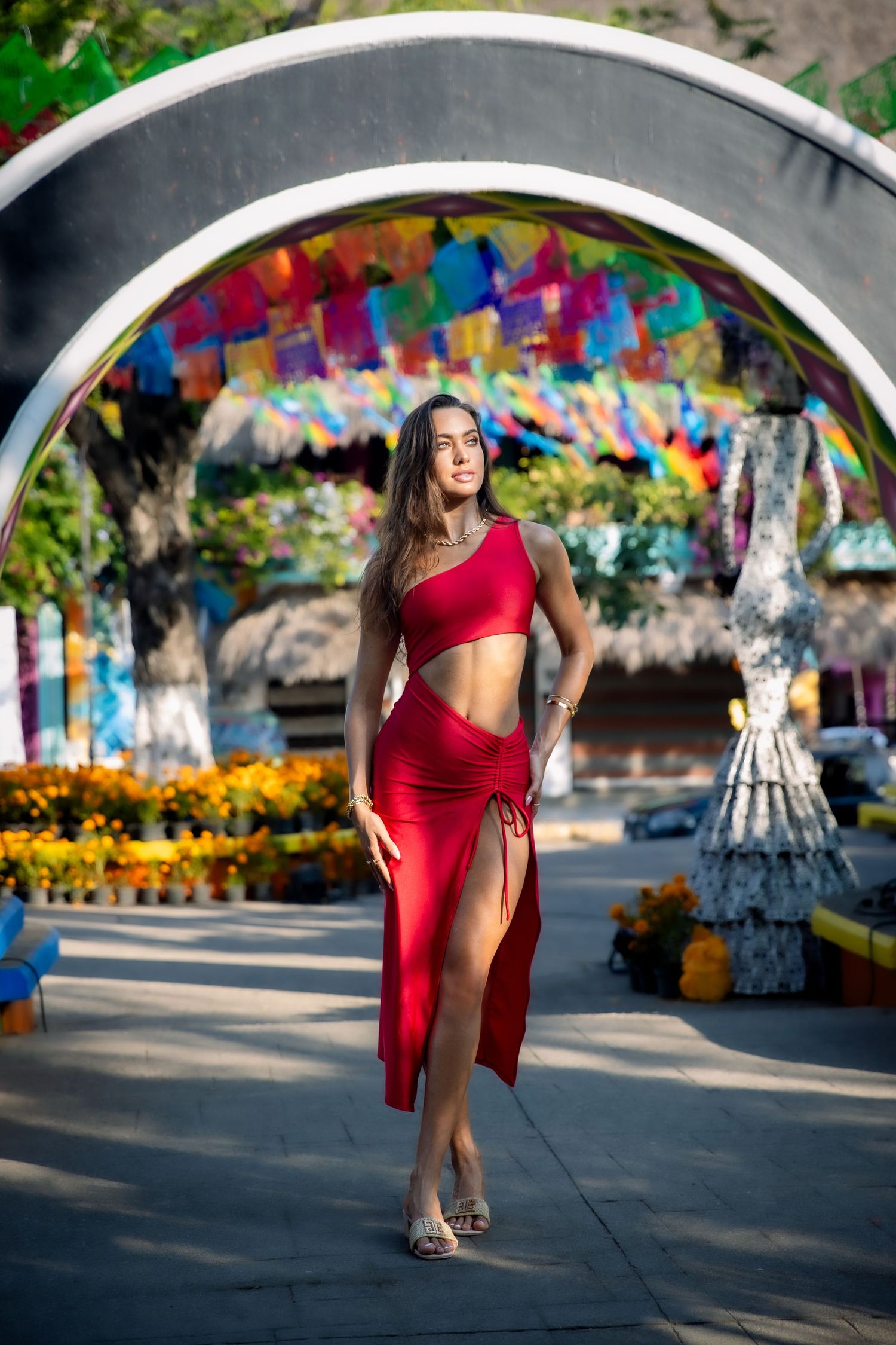 A woman in a red two-piece outfit walks through a colorful archway decorated with flowers and vibrant streamers.
