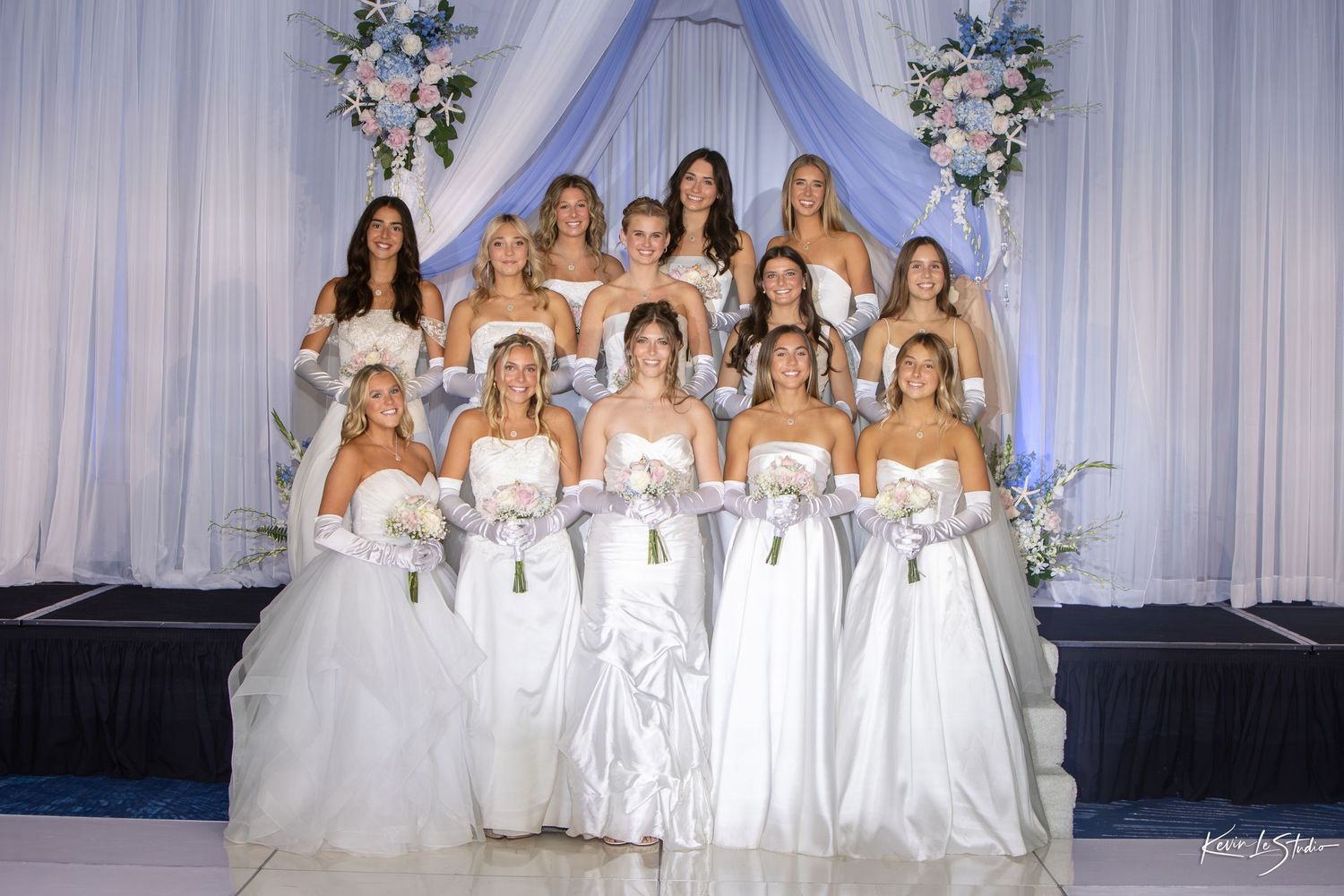 A group of bridesmaids in elegant white gowns pose together in front of a decorated backdrop with floral arrangements.