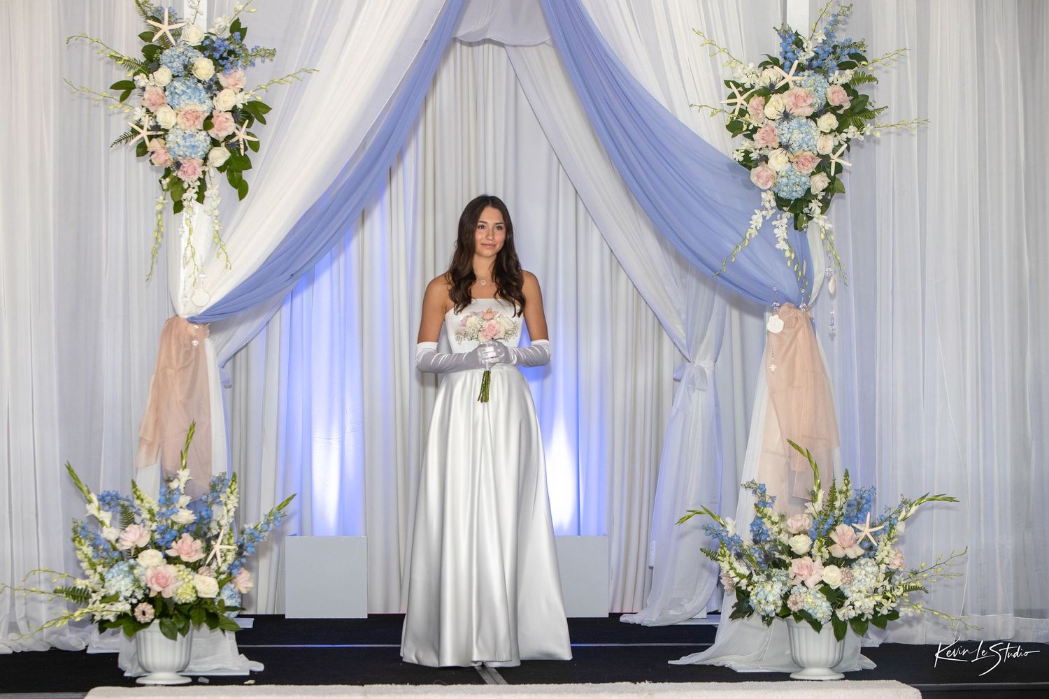 Elegant wedding ceremony setup with white draped fabric arch, blue uplighting and floral arrangements on pedestals.
