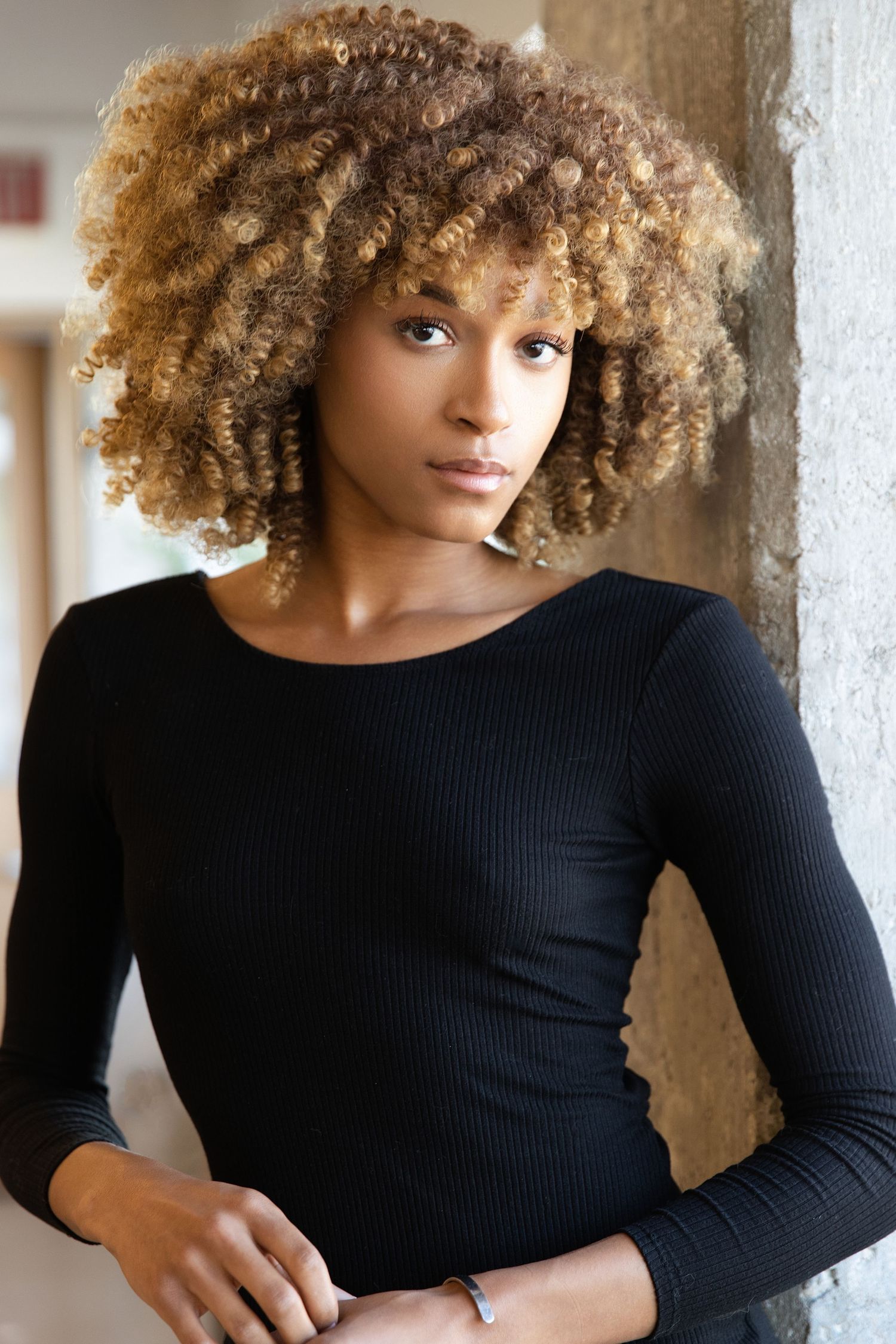 Person with voluminous curly blonde afro hair wearing a black long sleeve top poses against a light colored wall.