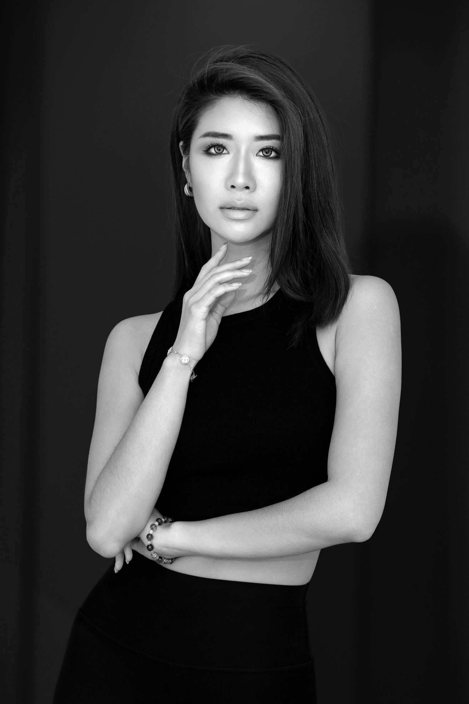 Black and white portrait of a woman in sleeveless dress posing elegantly with hand near face against dark background.