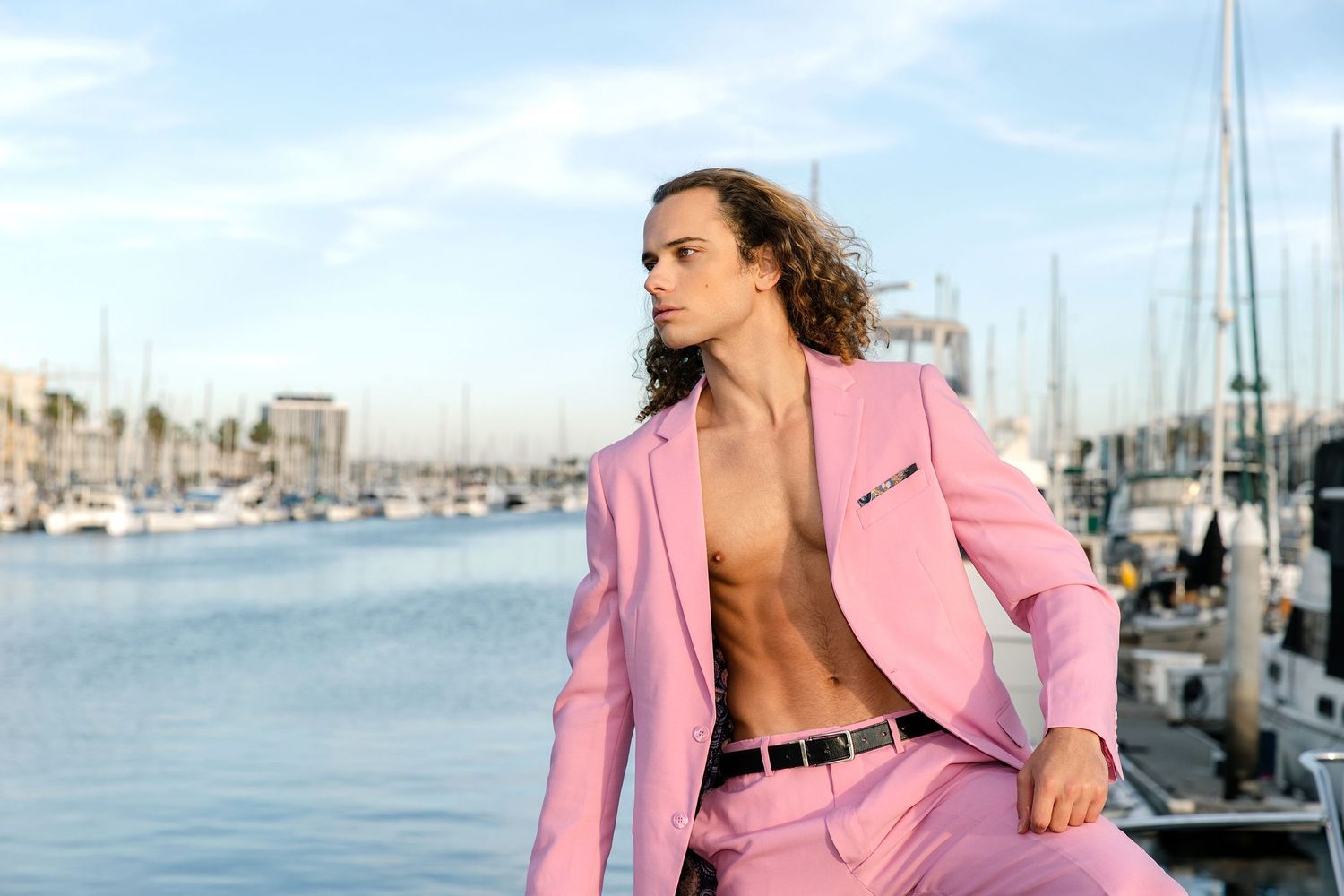 Woman in pink blazer poses on a marina dock with boats and waterfront buildings in the background.