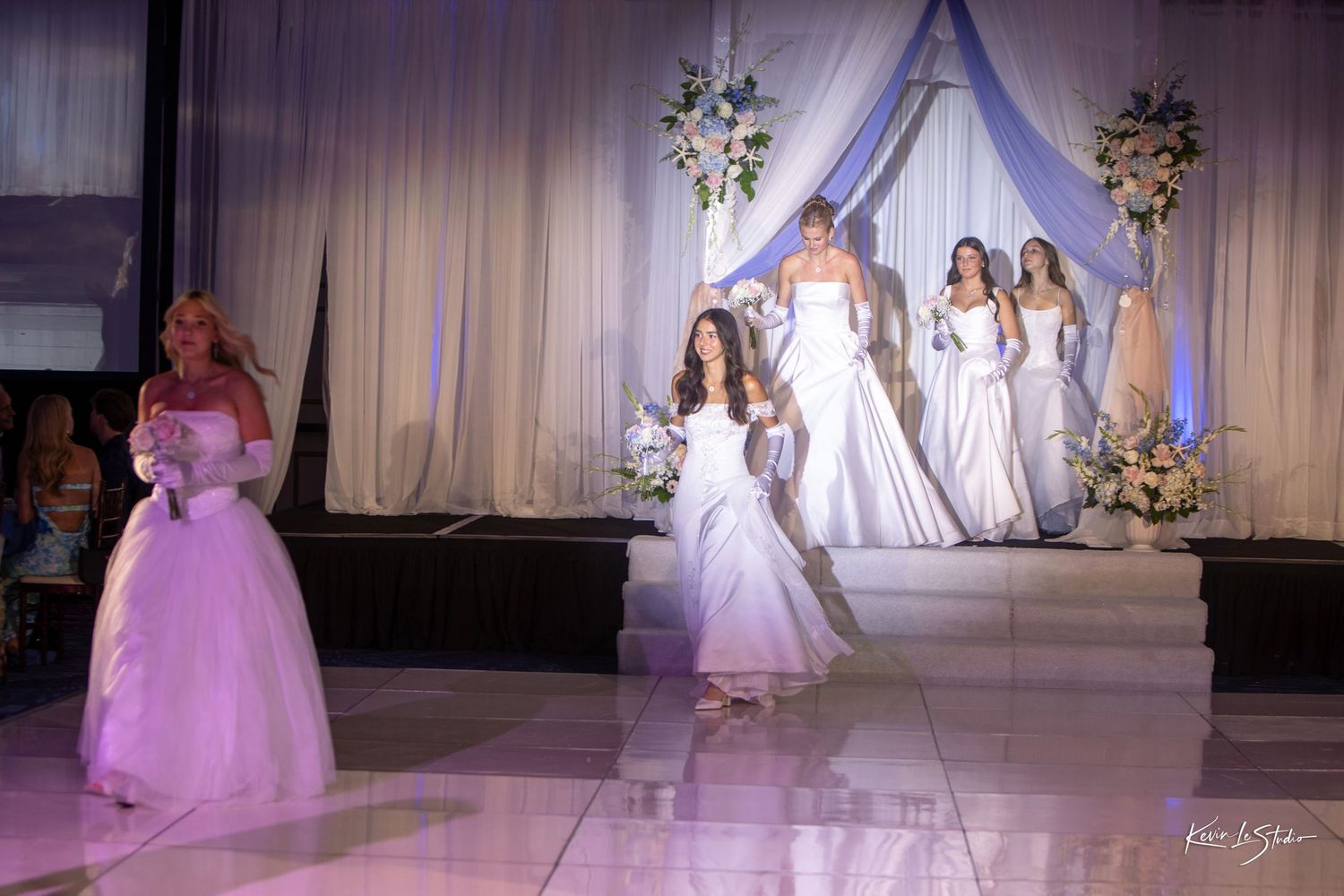 Models showcase elegant wedding gowns and formal dresses on a bridal fashion show runway with draped backdrop and floral decor.