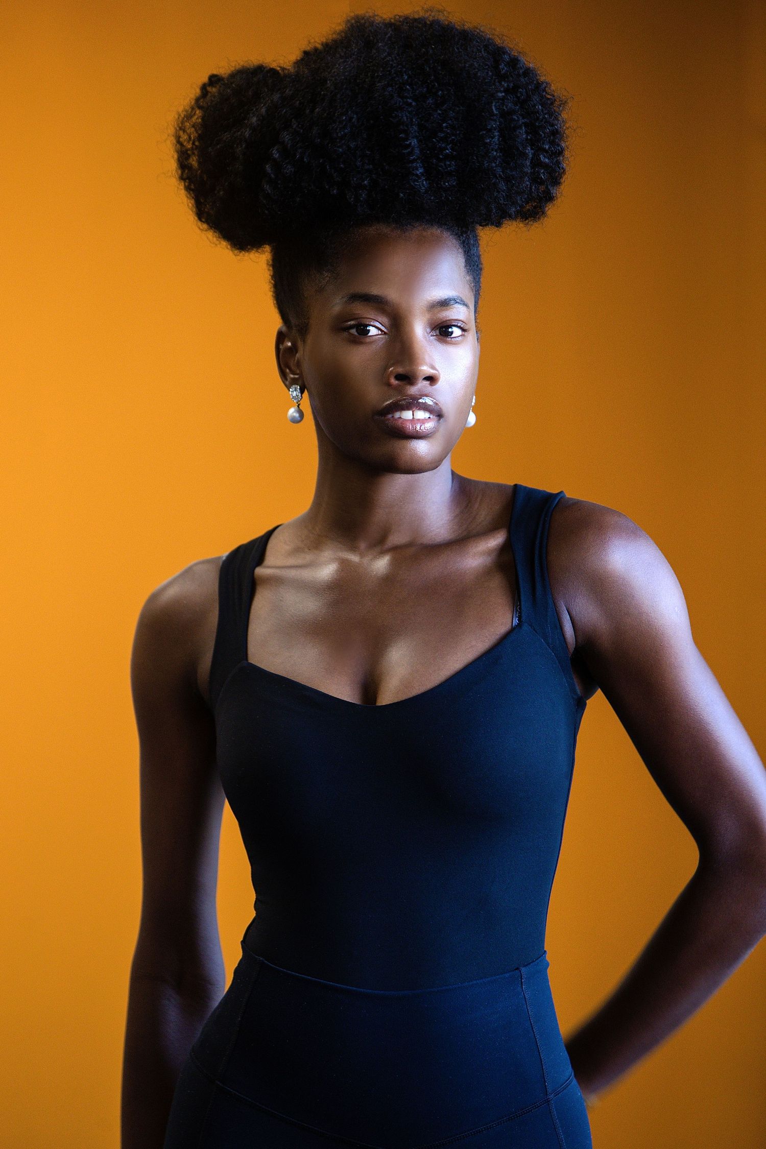 Portrait against orange background showing natural afro hairstyle and black sleeveless dress in studio lighting.