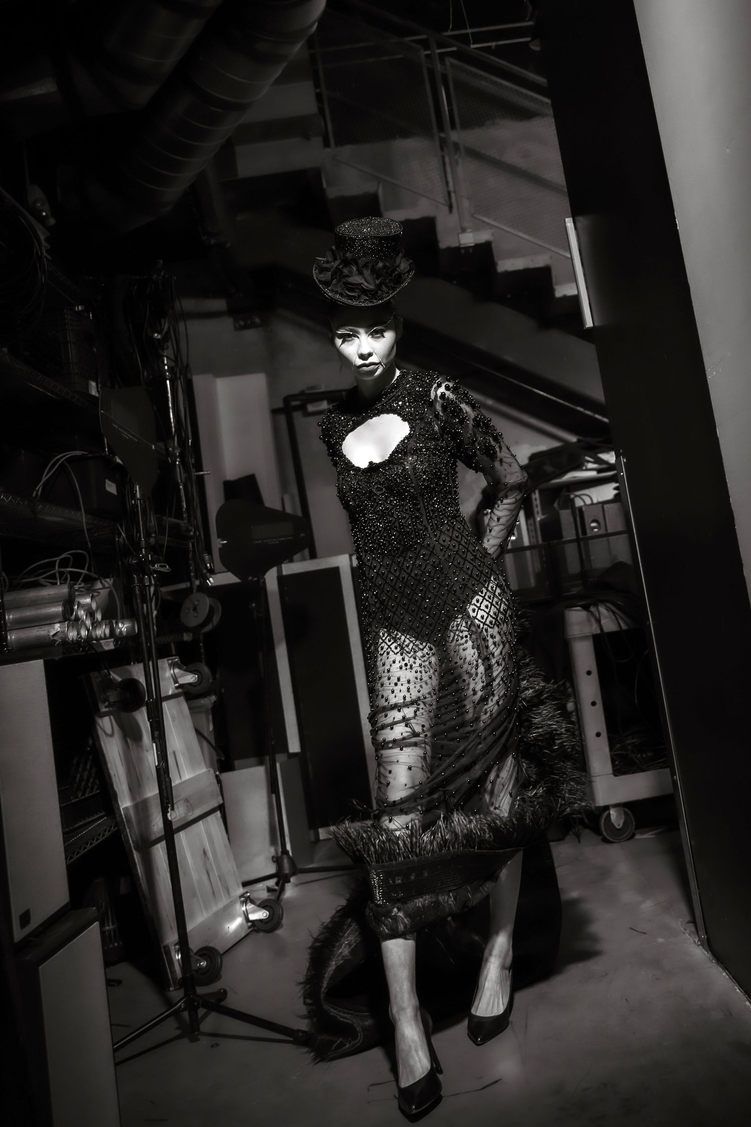 A dramatic black and white photo shows a figure in a fitted lace dress posing in a dark stairwell with theatrical lighting.