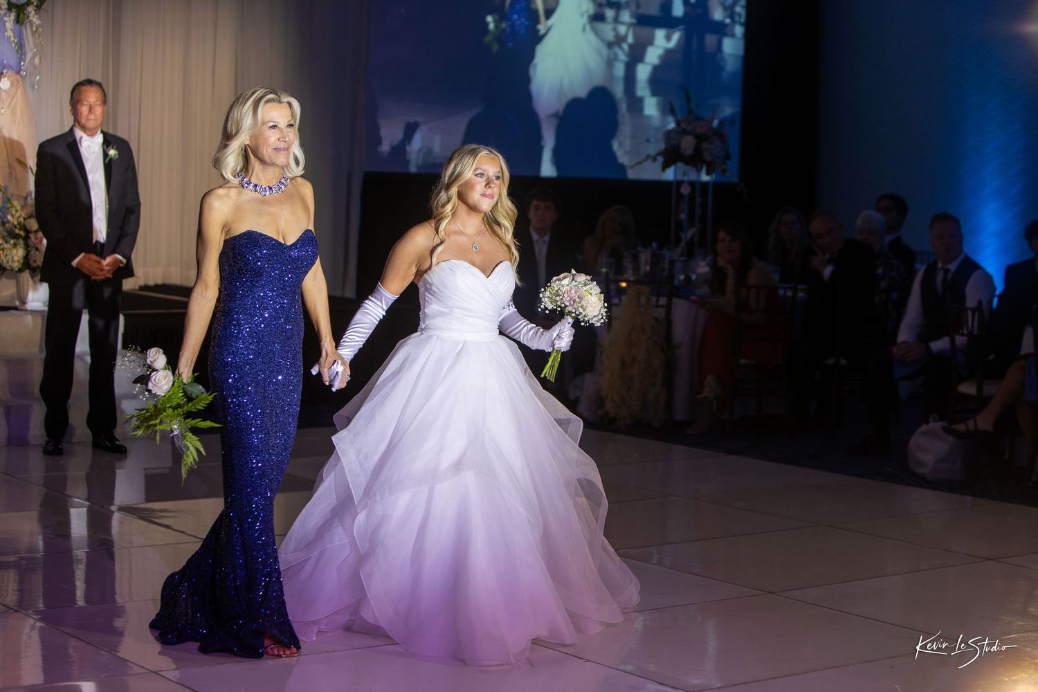 A bride in a white ballgown walks alongside a woman in a navy blue dress during an elegant wedding reception.