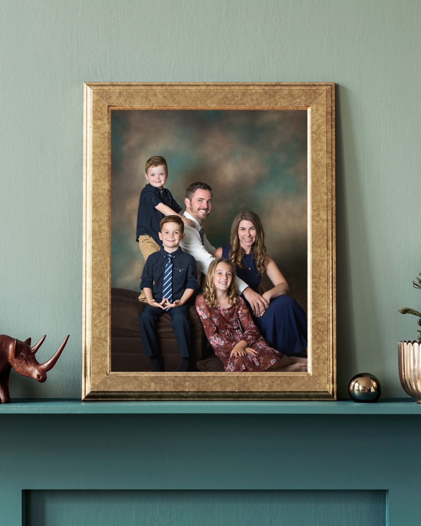 A framed family portrait in a gold frame displayed on a teal mantle, featuring parents and two children posing together.