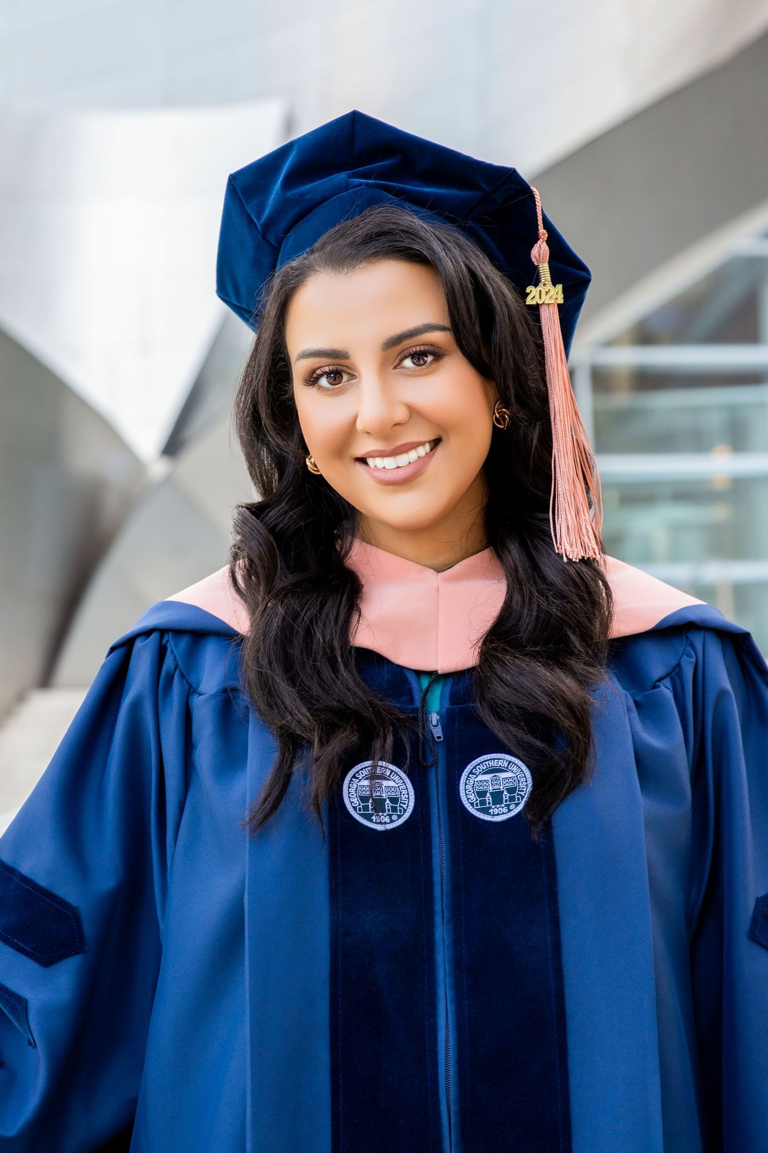 Young woman smiling in blue doctoral graduation gown and cap with pink tassel and hood at outdoor ceremony.