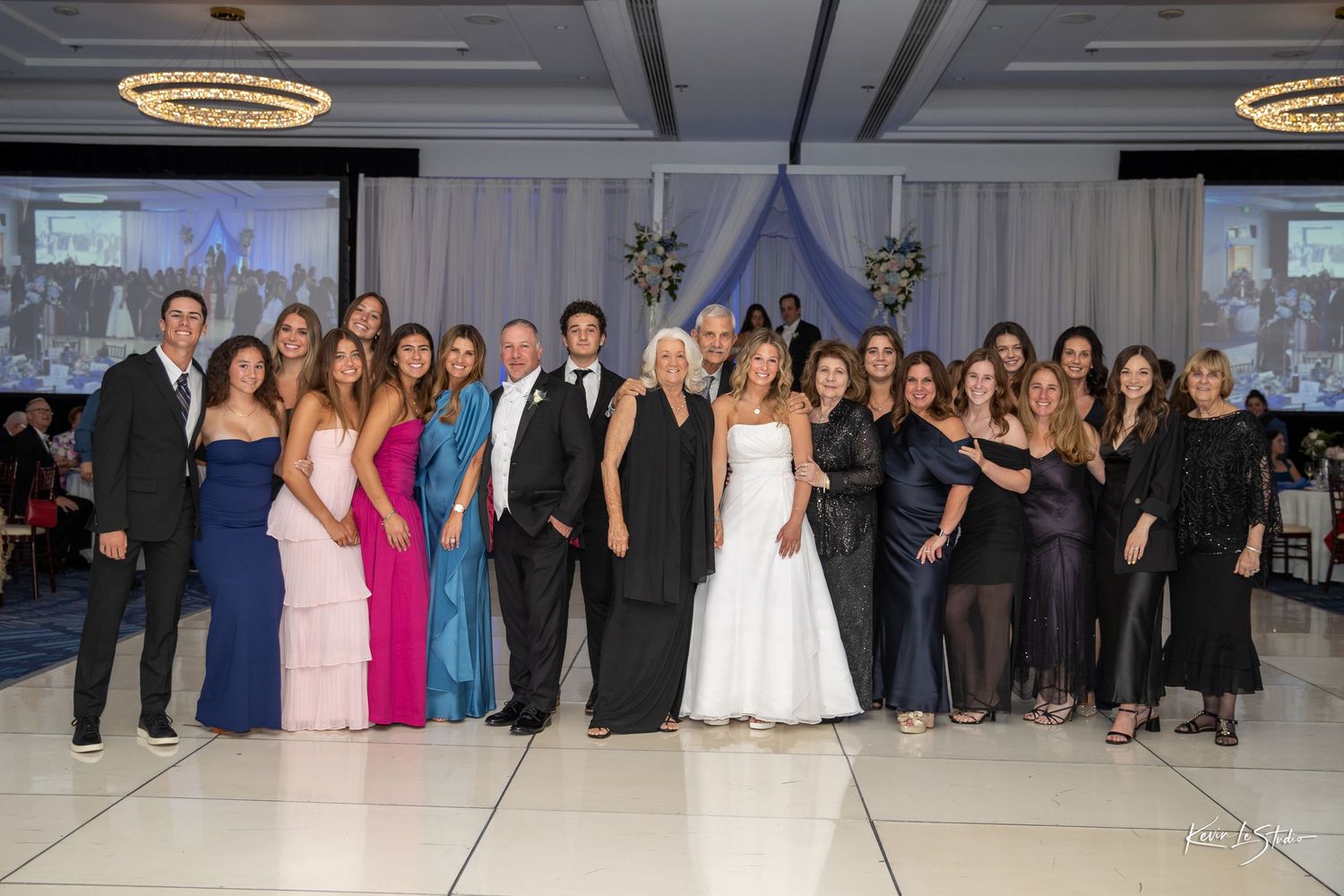 Group of wedding guests and party members posing together on a dance floor with circular chandeliers overhead.