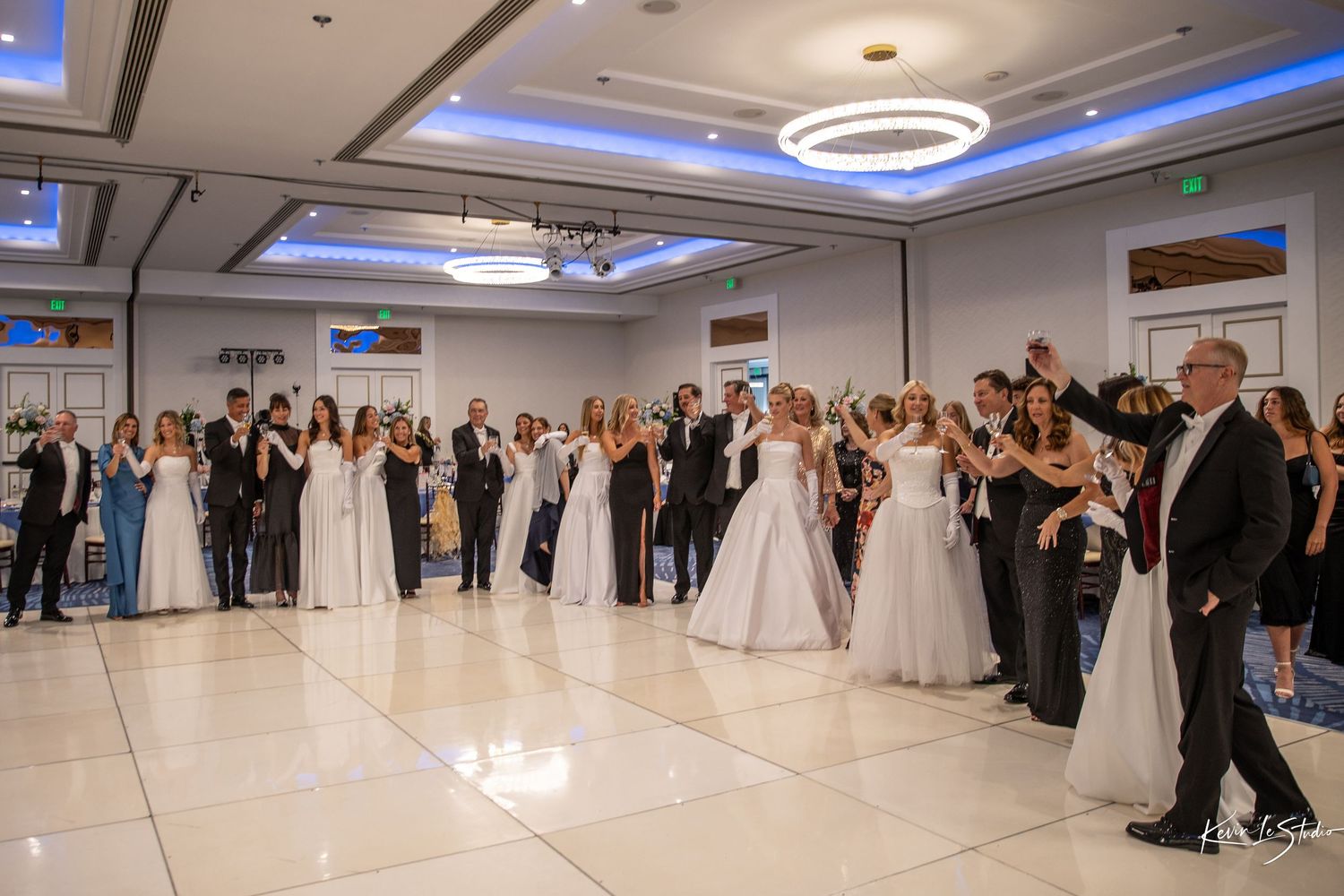 Wedding reception guests in formal attire gather on a white dance floor under blue-lit circular ceiling fixtures.