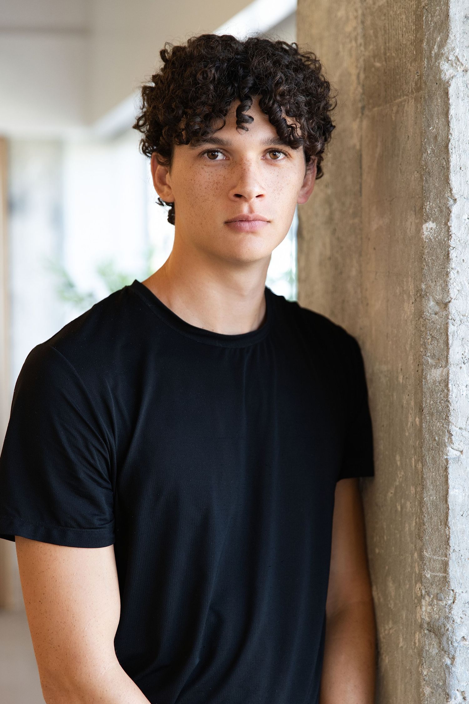 A person with curly dark hair wearing a black t-shirt poses against a light colored wall in a series of similar shots.