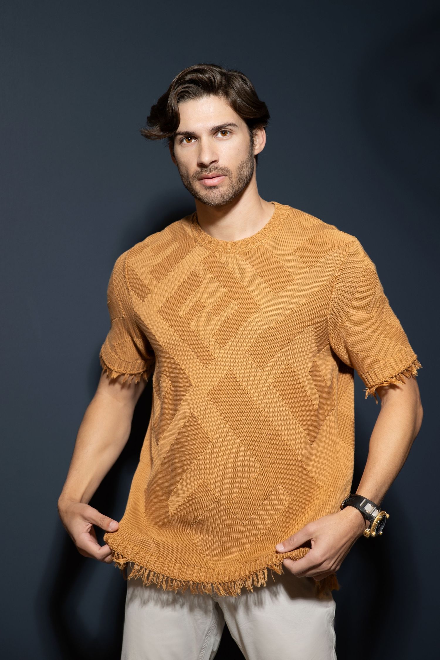 A person wearing a tan knitted sweater with geometric patterns and frayed edges poses against a dark background.