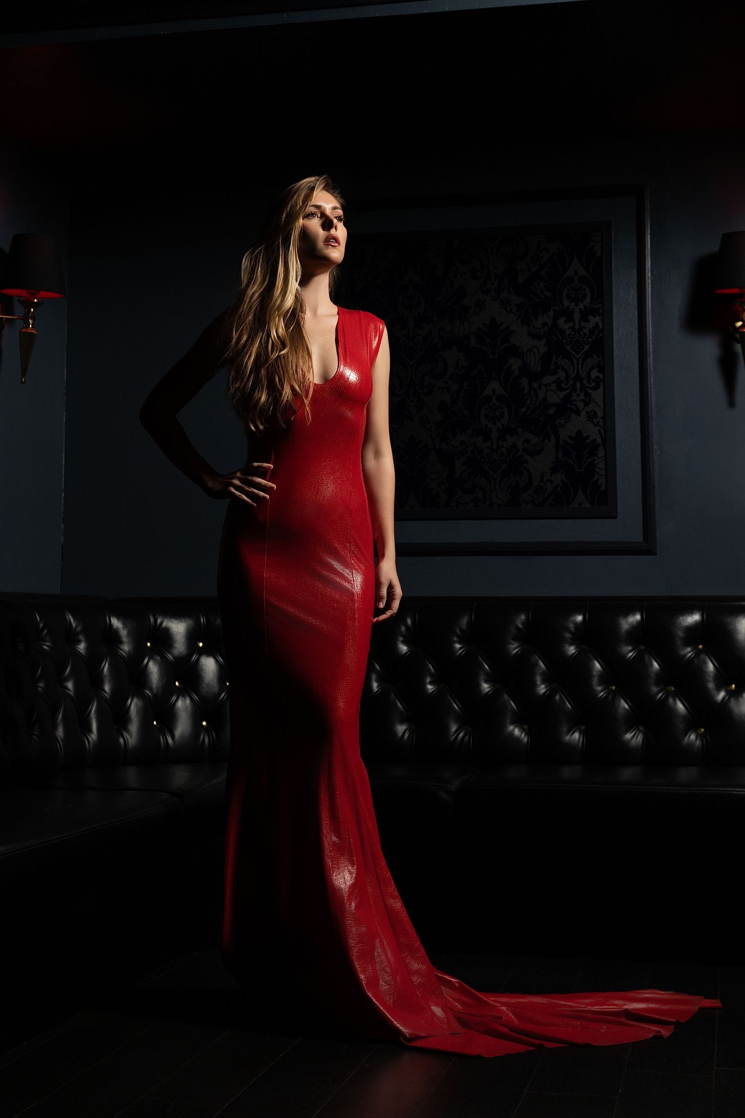 A dramatic fashion photo of a long red evening gown with a flowing train against a dark background with tufted seating.