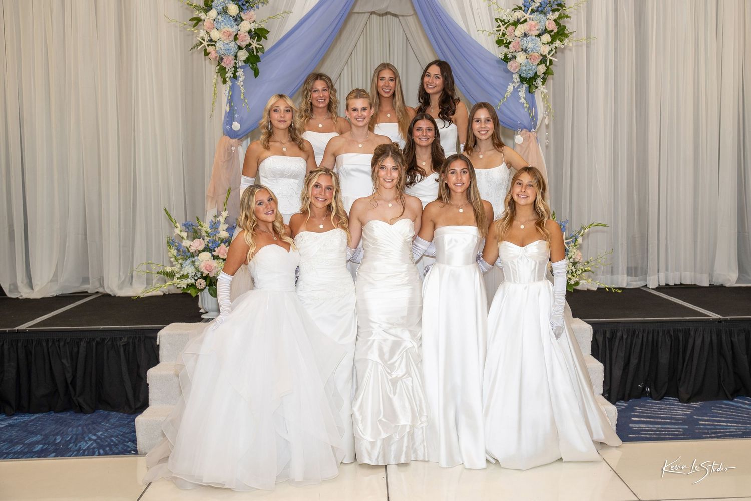 A group of women in white dresses pose together in front of a floral arch at an elegant formal event.