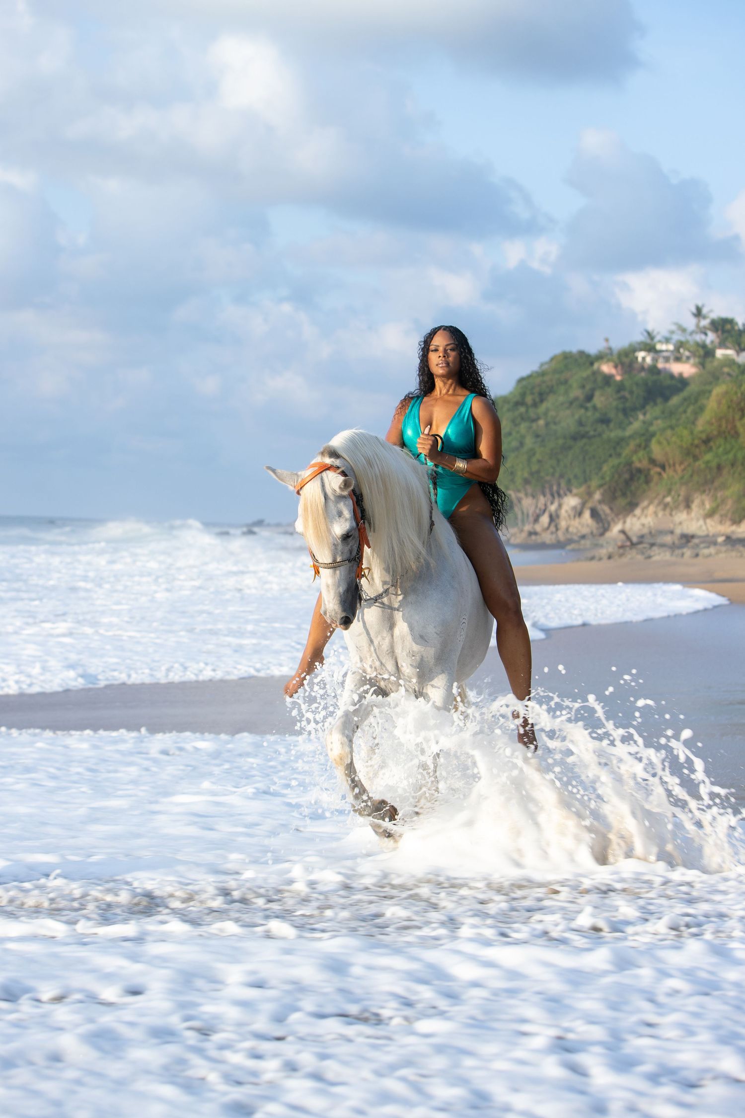 A person rides a white horse through crashing ocean waves along a scenic beach with cliffs in the background.