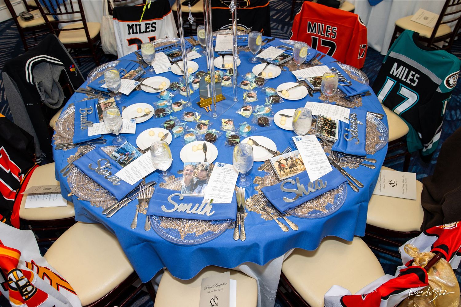 Multiple round tables with blue tablecloths and place settings arranged in a banquet hall setting for an event.
