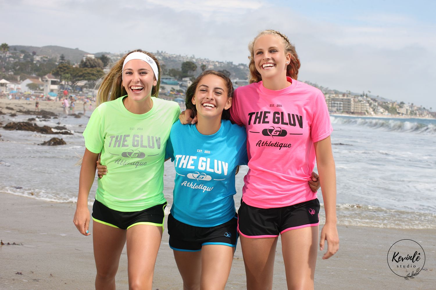 Three runners wearing bright neon workout shirts laugh together while exercising on a scenic beach.