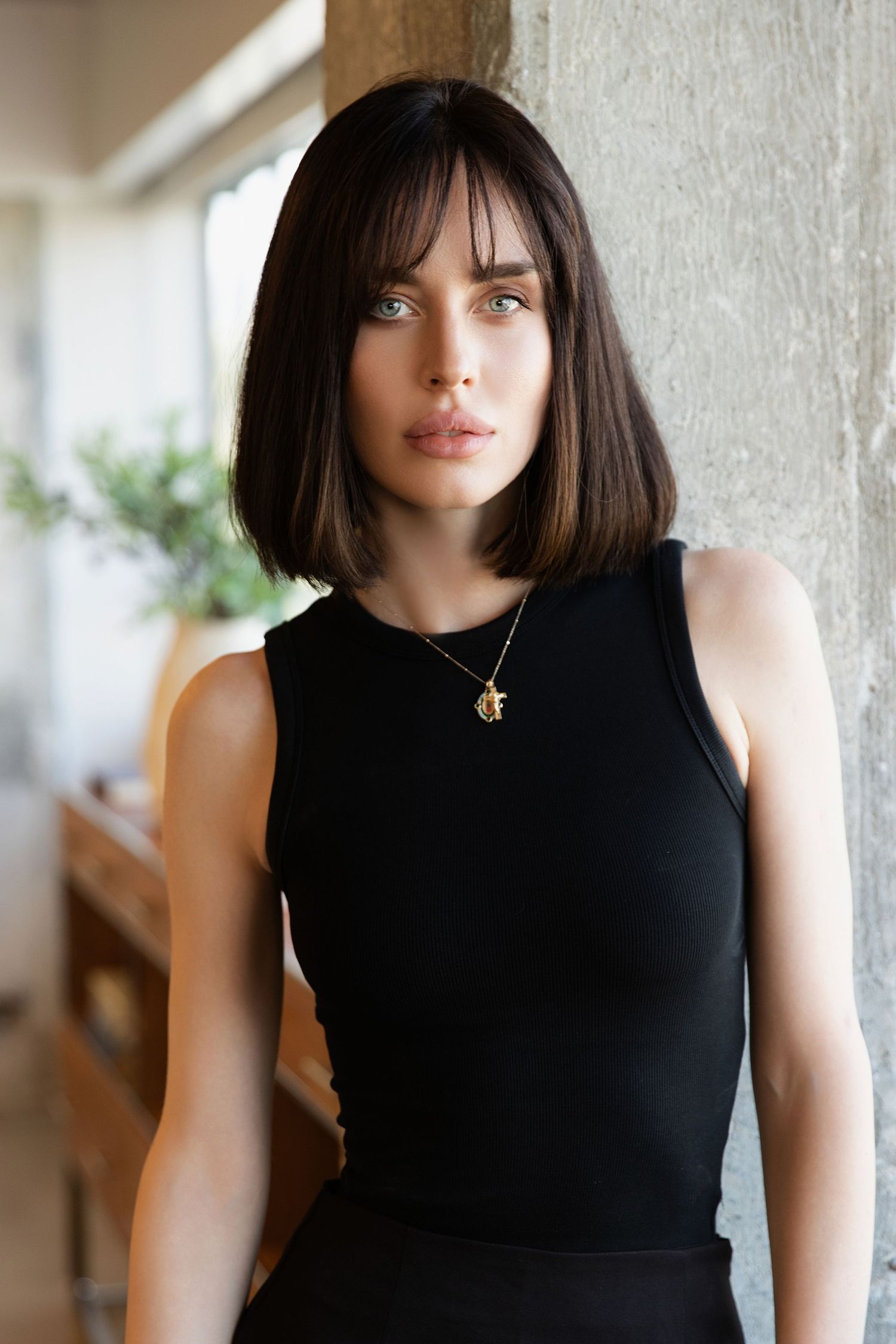 Portrait of someone with dark straight bob haircut wearing a black sleeveless top and delicate pendant necklace against light wall.