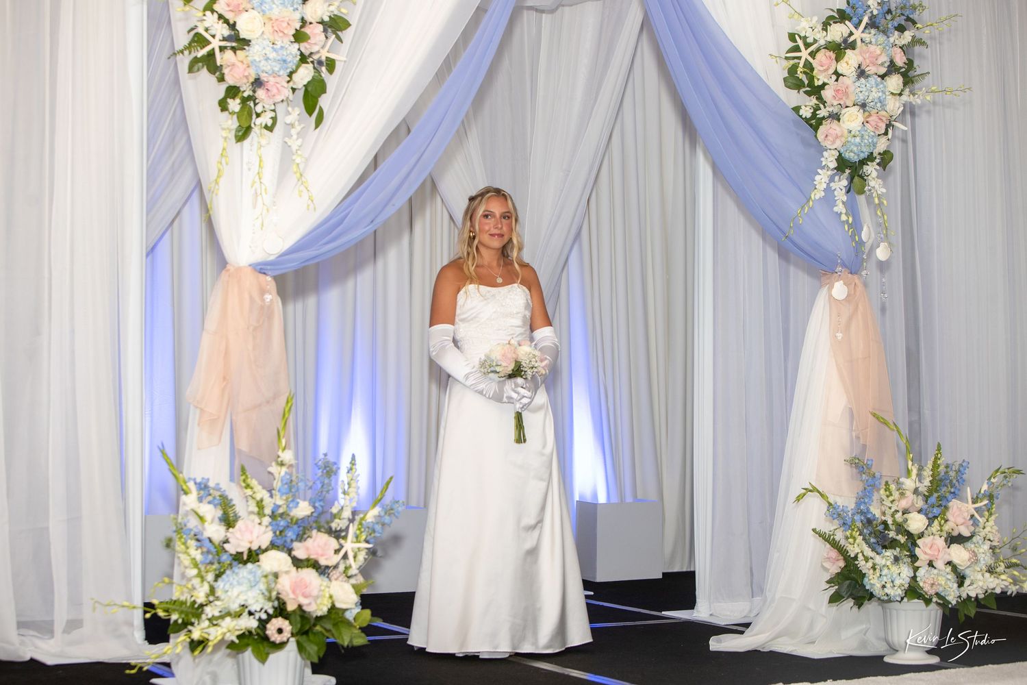 Elegant white wedding backdrop with flowing fabric drapes, floral arrangements, and soft blue uplighting creates a romantic atmosphere.