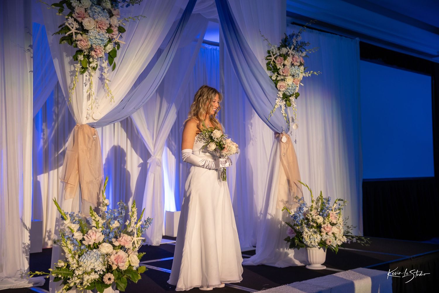 Elegant wedding ceremony setup with white draping, floral arrangements and blue lighting creating a romantic atmosphere.