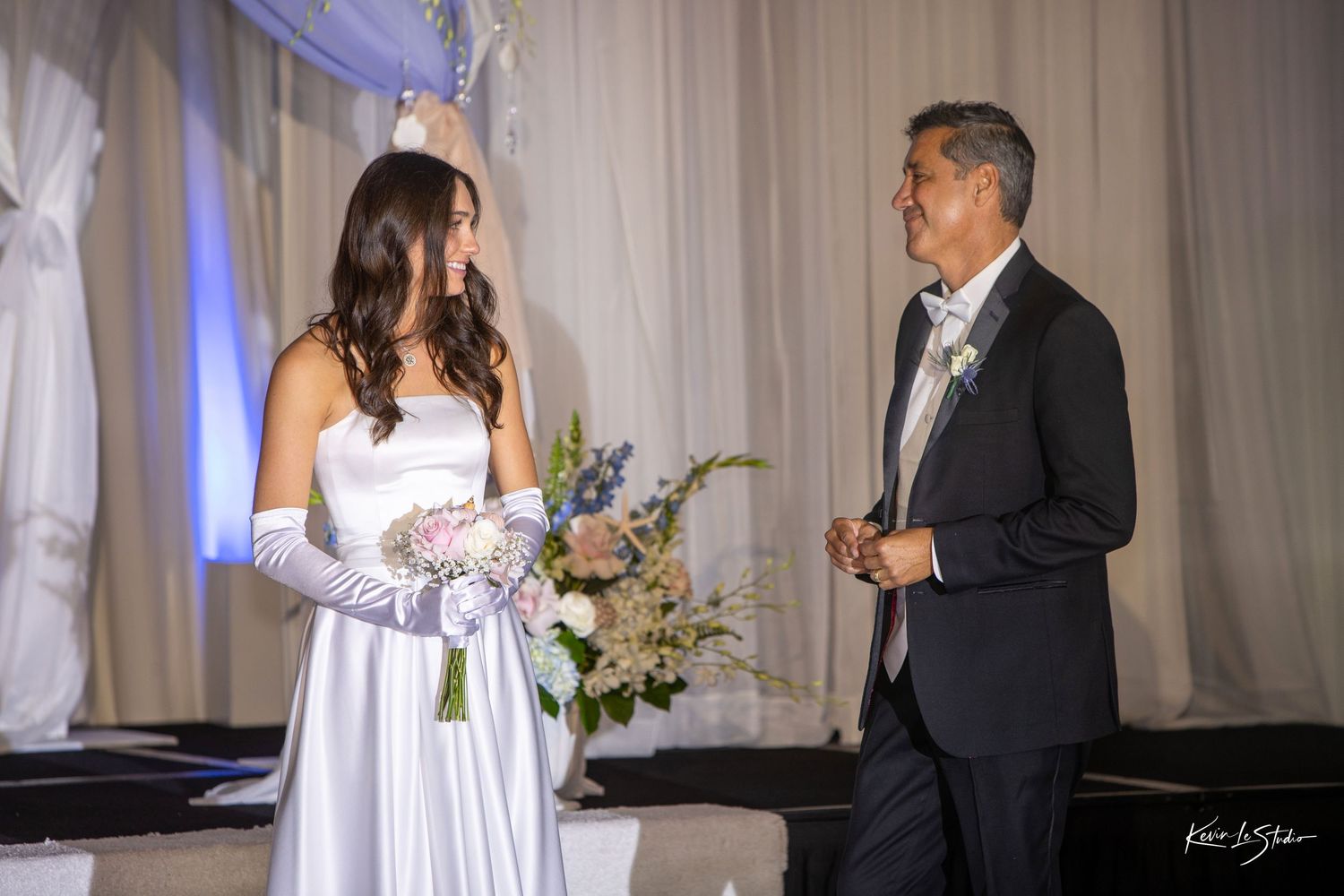 A couple shares a tender moment during their wedding ceremony in an elegant white-draped venue with soft lighting.
