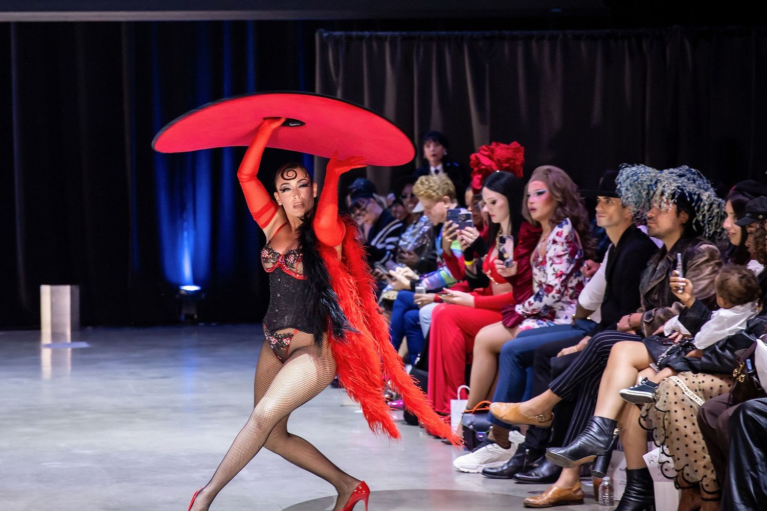 A dancer performs with a flowing red fabric on a runway while audience members watch from the sideline.