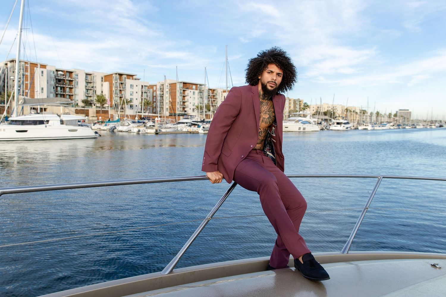 A person in a burgundy suit poses on a yacht with a scenic harbor and buildings in the background.