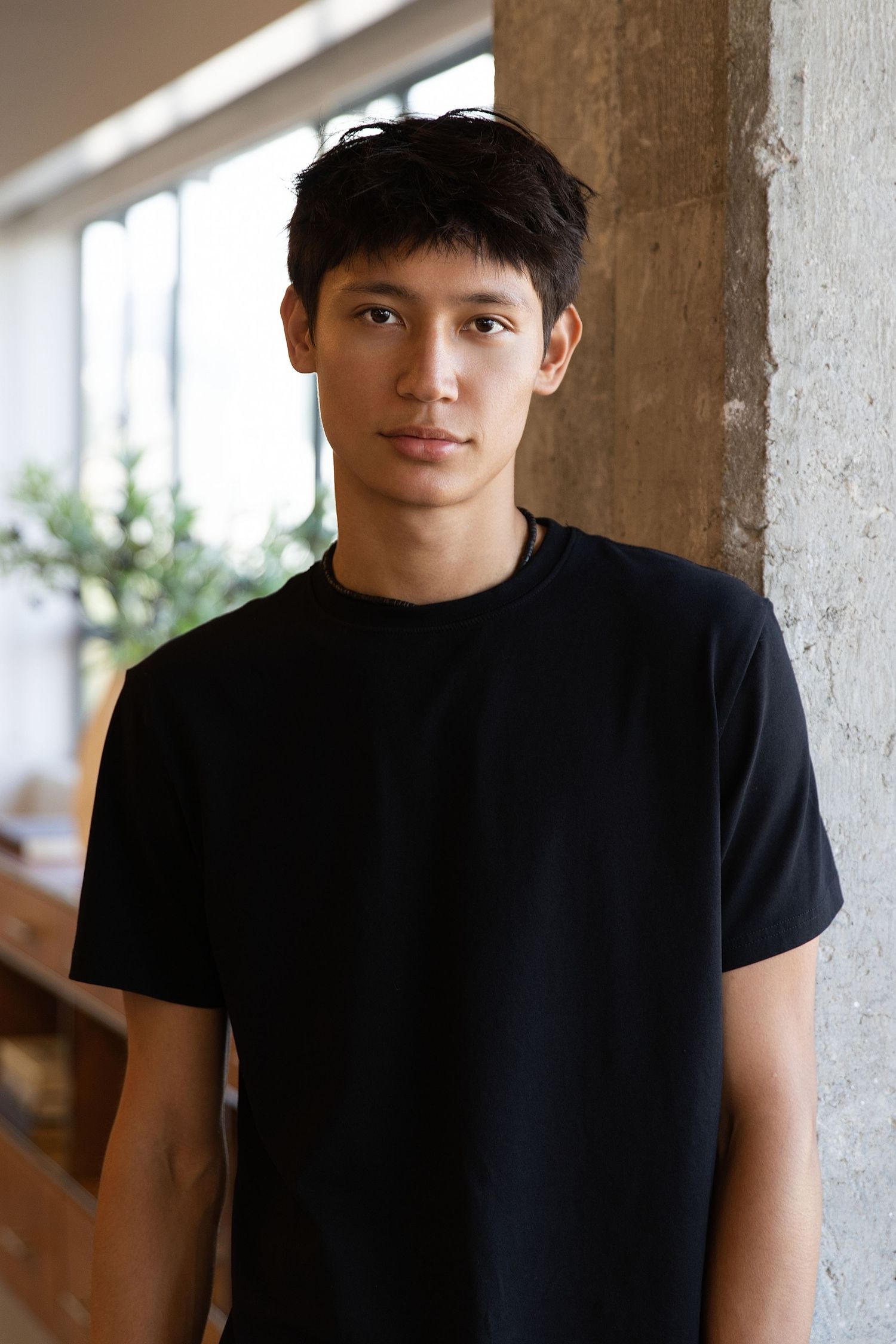 Person wearing a black t-shirt and cap standing against a light-colored wall.
