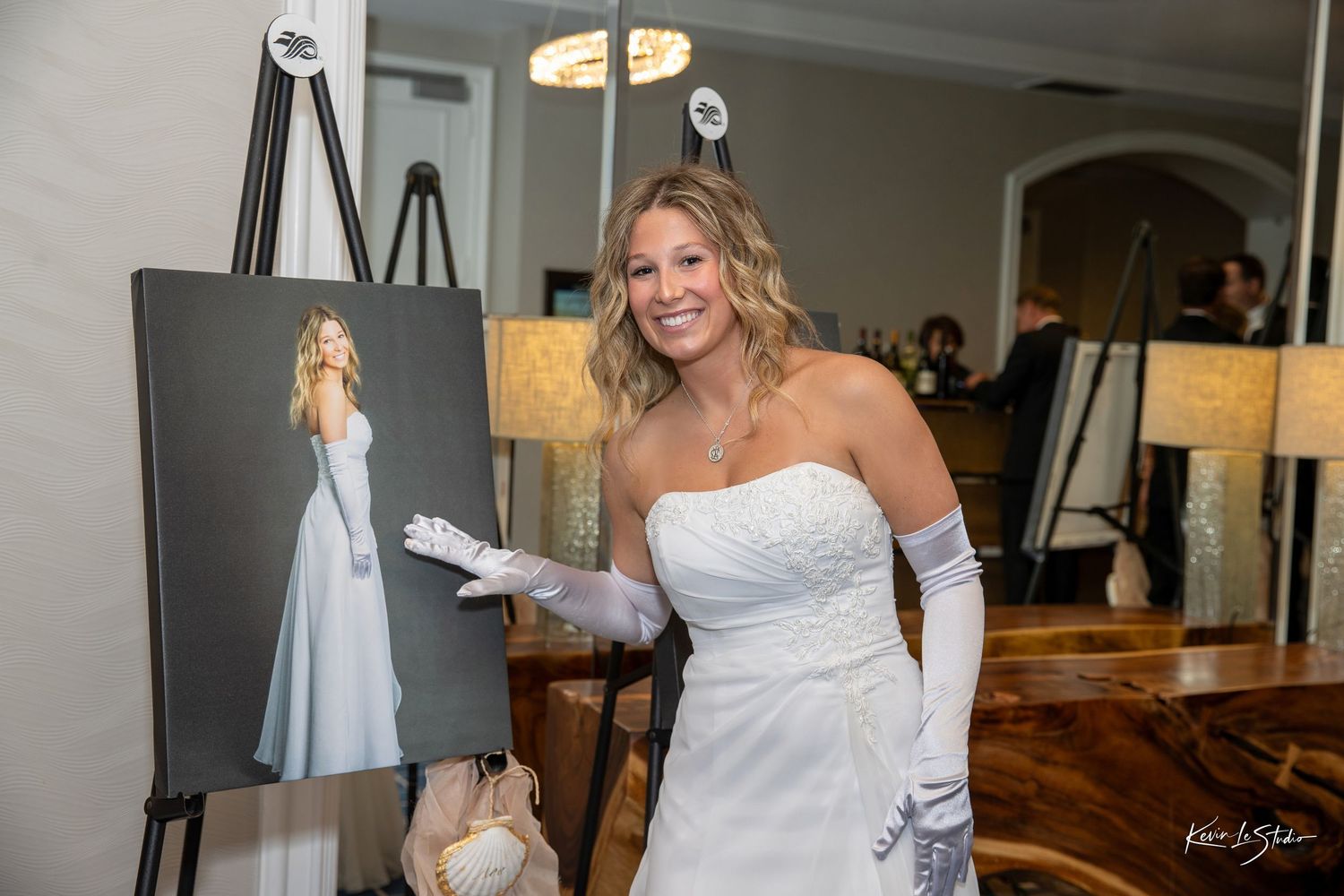 A person in a white wedding dress and gloves stands next to an easel displaying artwork in an elegant room.