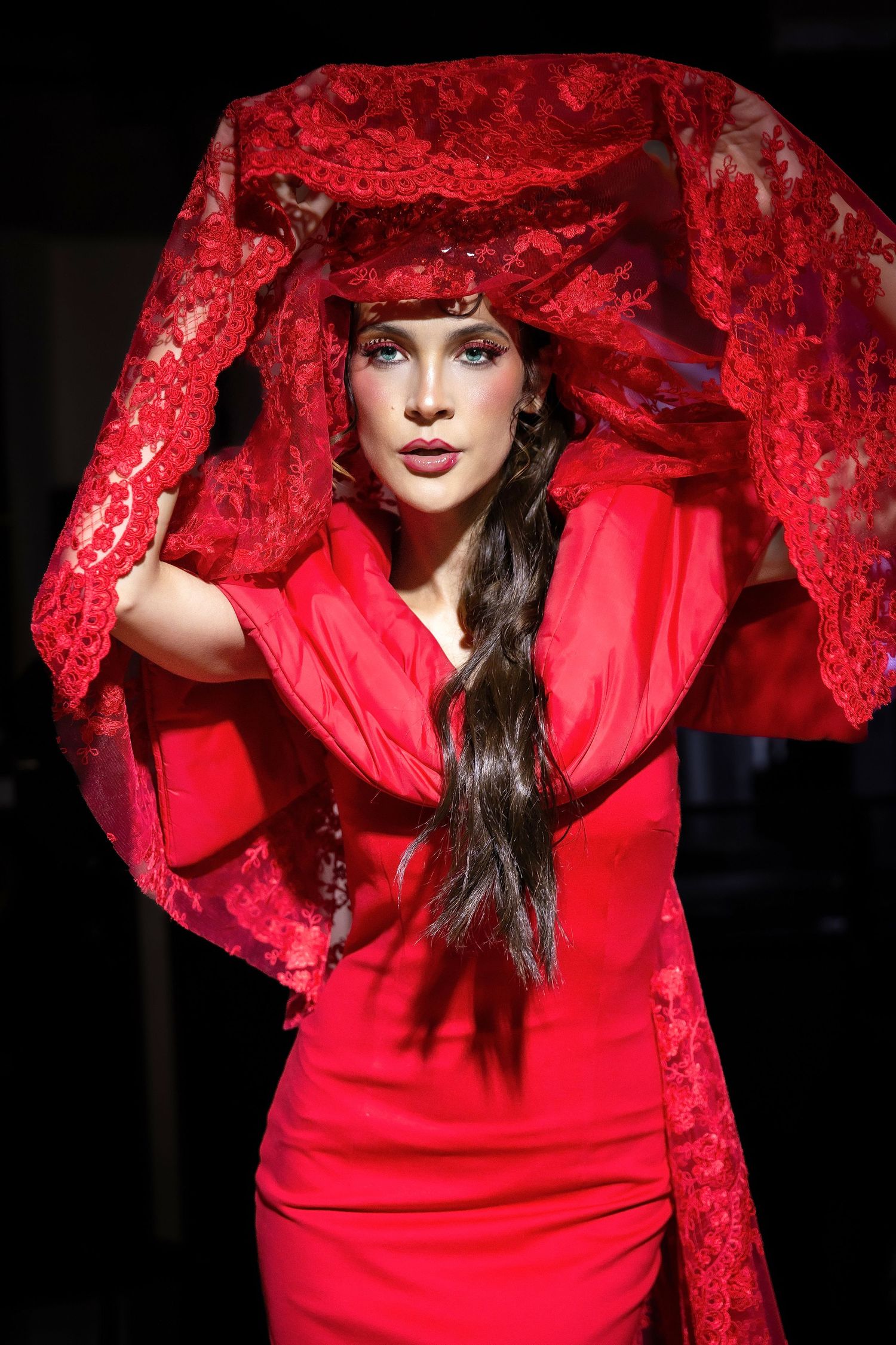 Fashion model wearing a dramatic red ensemble with lace detailing walks the runway against dark background.