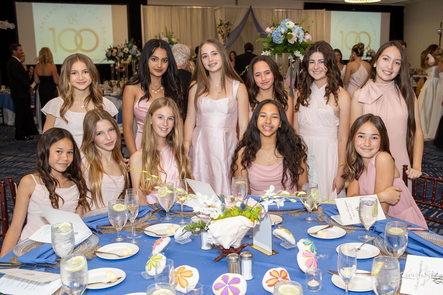 A group of guests in pink and white dresses gather around an elegantly decorated table at a formal event.