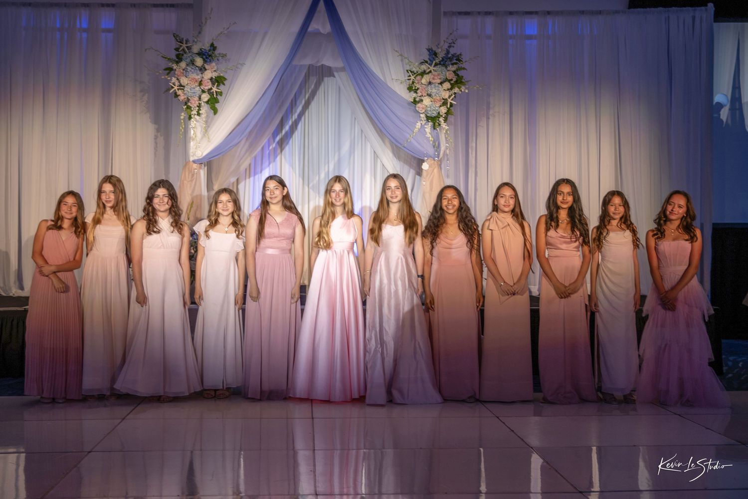 A row of bridesmaids in blush pink dresses stand in front of an elegant white draped backdrop with floral arrangements.