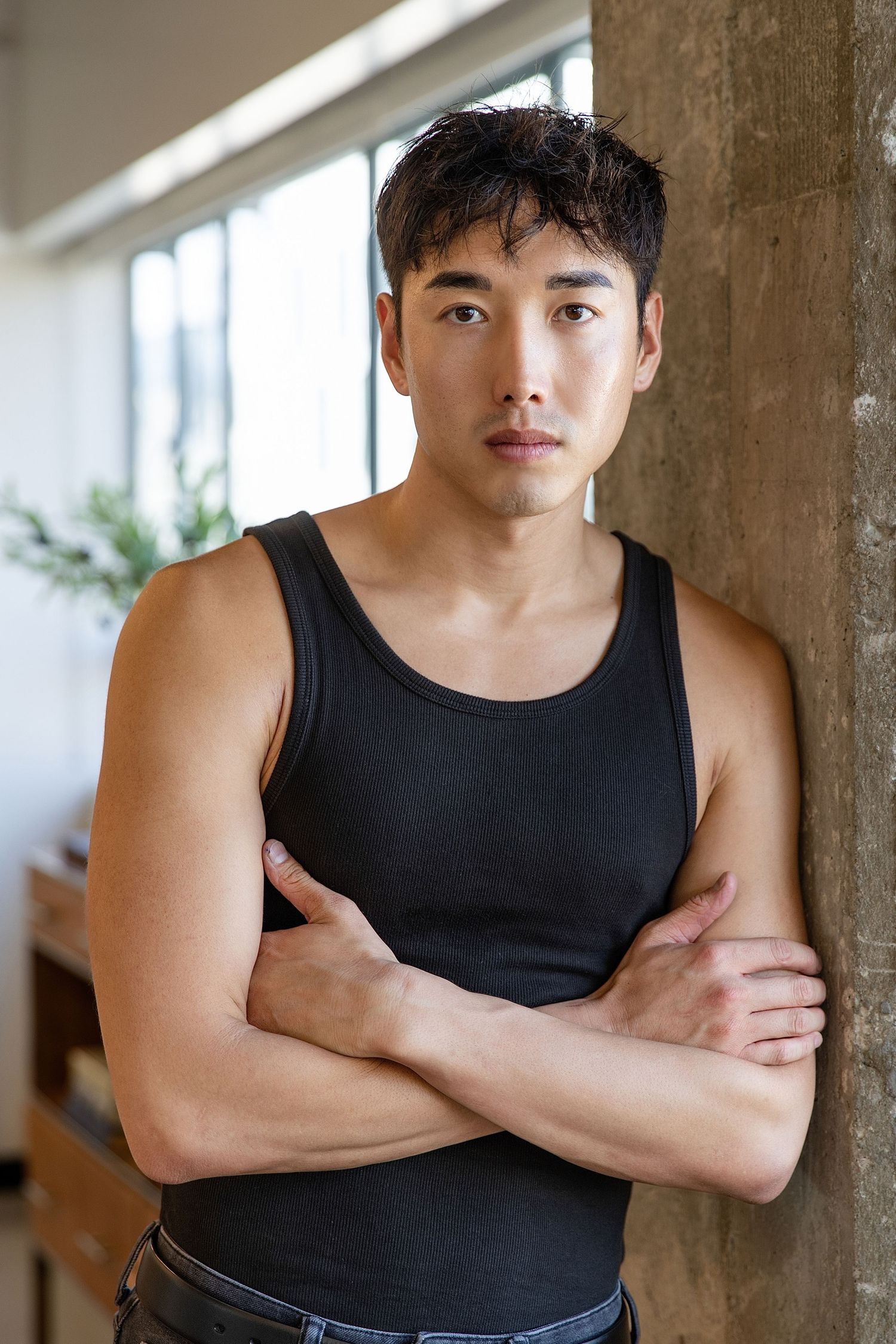 Young athletic person in black tank top standing with arms crossed against wall in natural lighting.