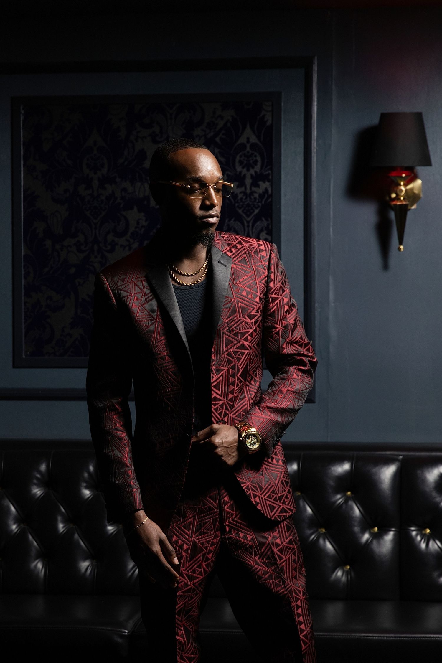A person in a stylish red patterned blazer poses against a dark leather couch in moody lighting.