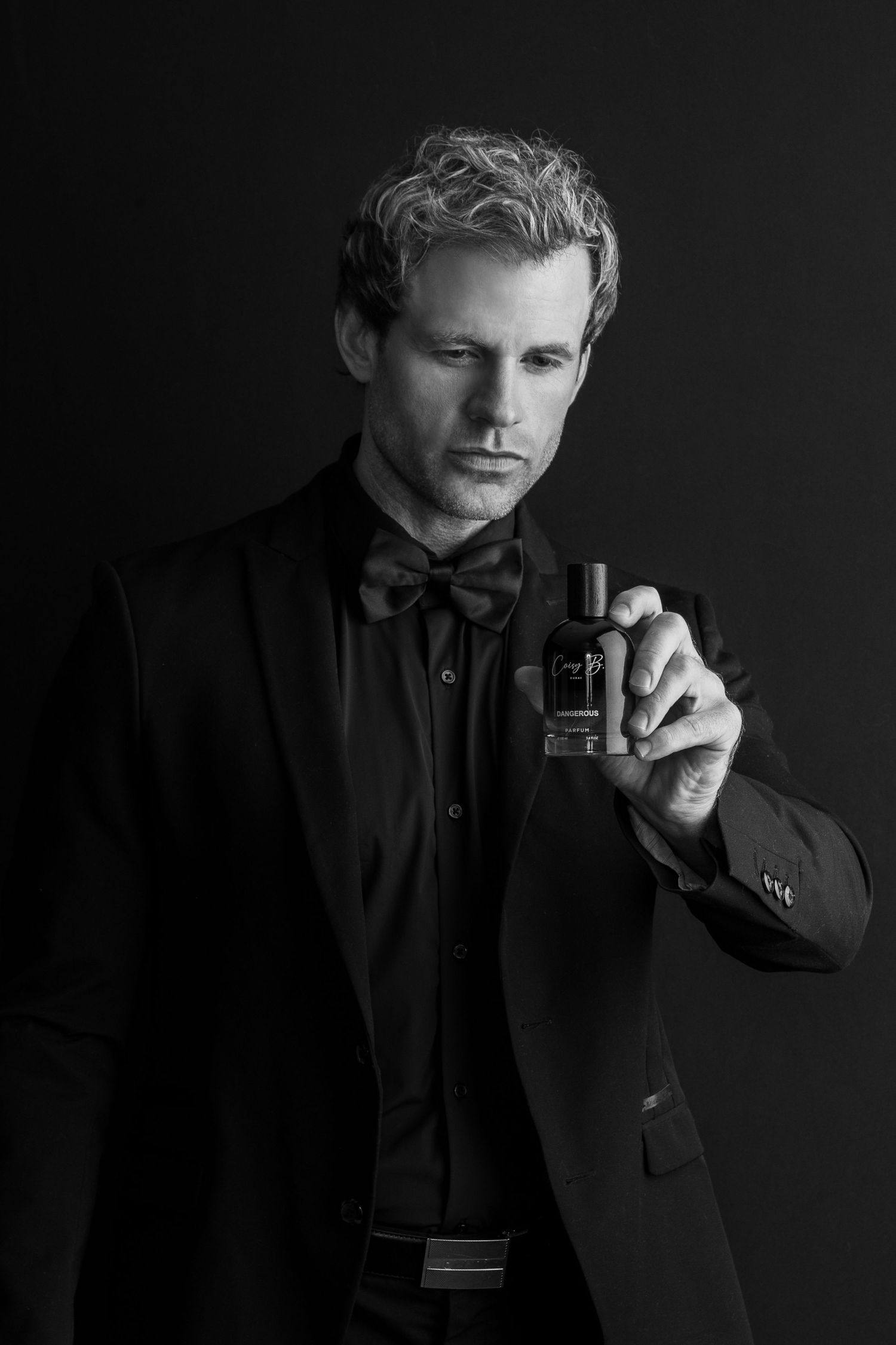 Elegant black and white portrait of person in tuxedo and bow tie holding cologne bottle against dark background.