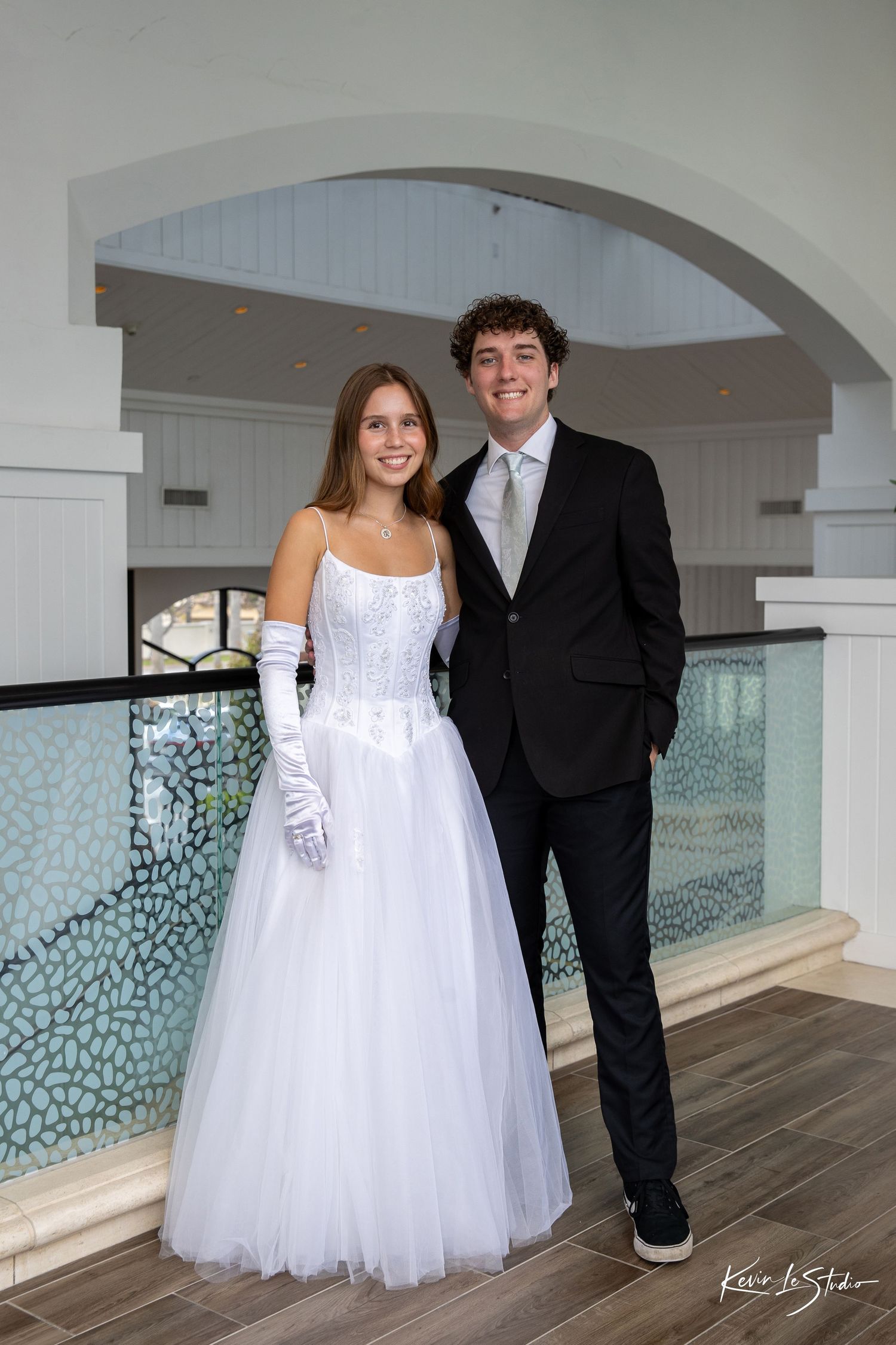 A couple in wedding attire poses together on a wooden deck with a decorative glass wall backdrop.