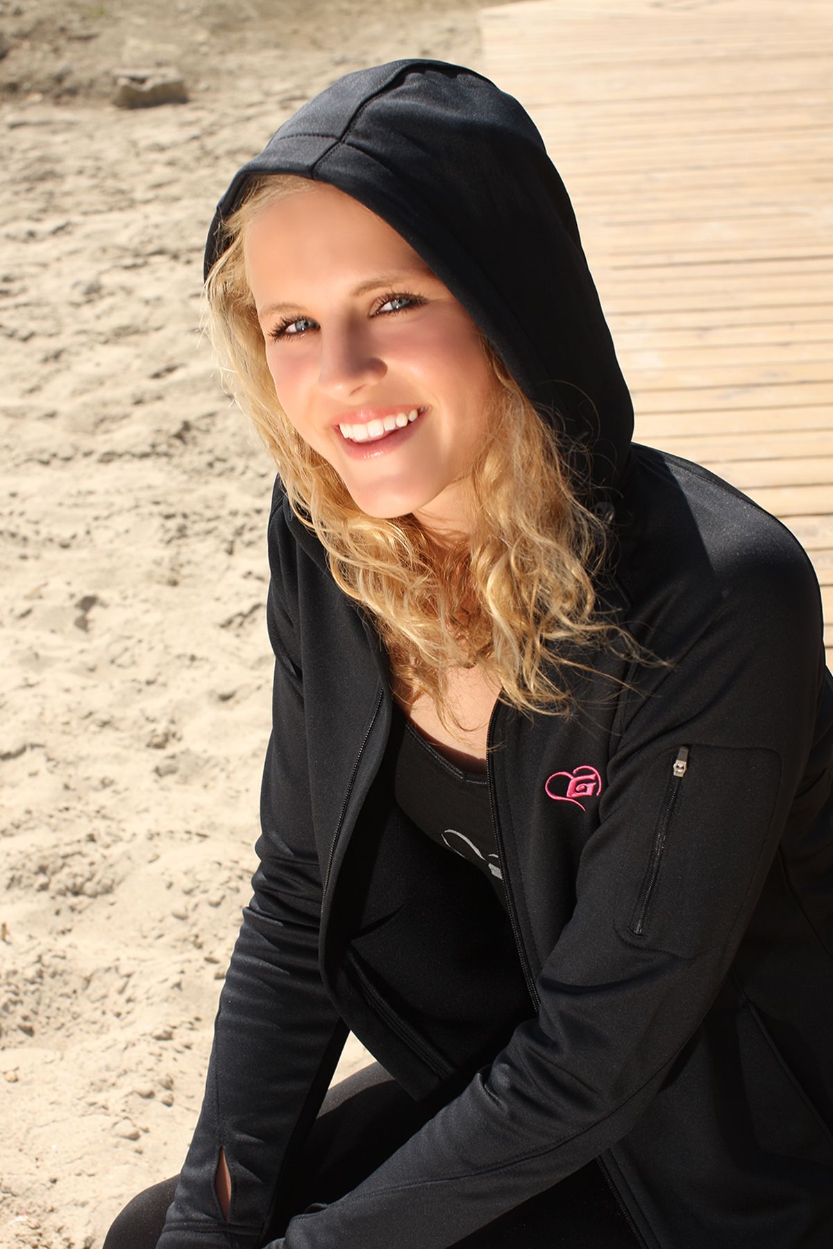 Woman wearing a black hooded jacket smiles while sitting outdoors on a sunny day.
