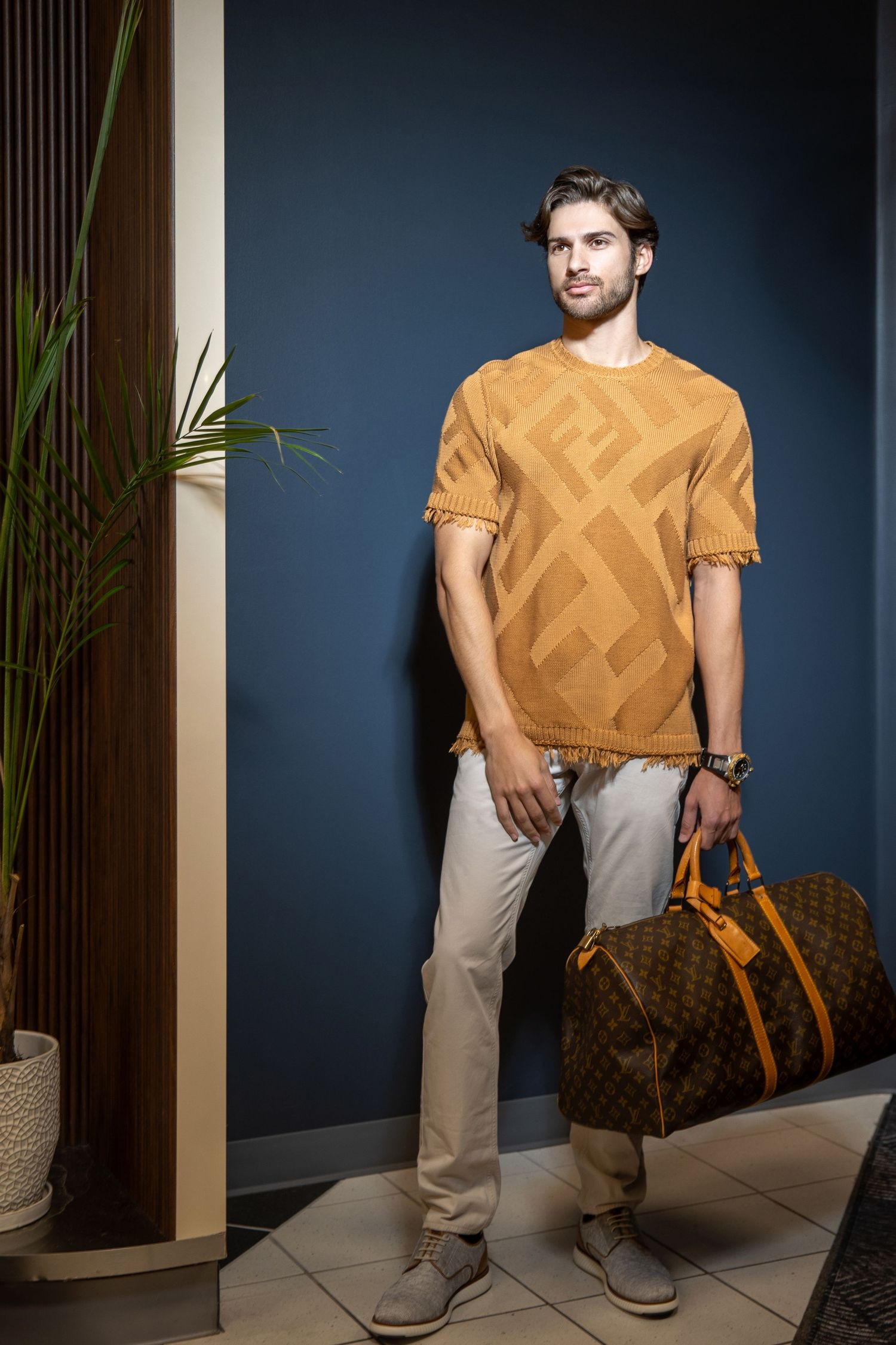 Fashionable outfit featuring mustard geometric t-shirt, beige pants and designer travel bag against dark blue wall.