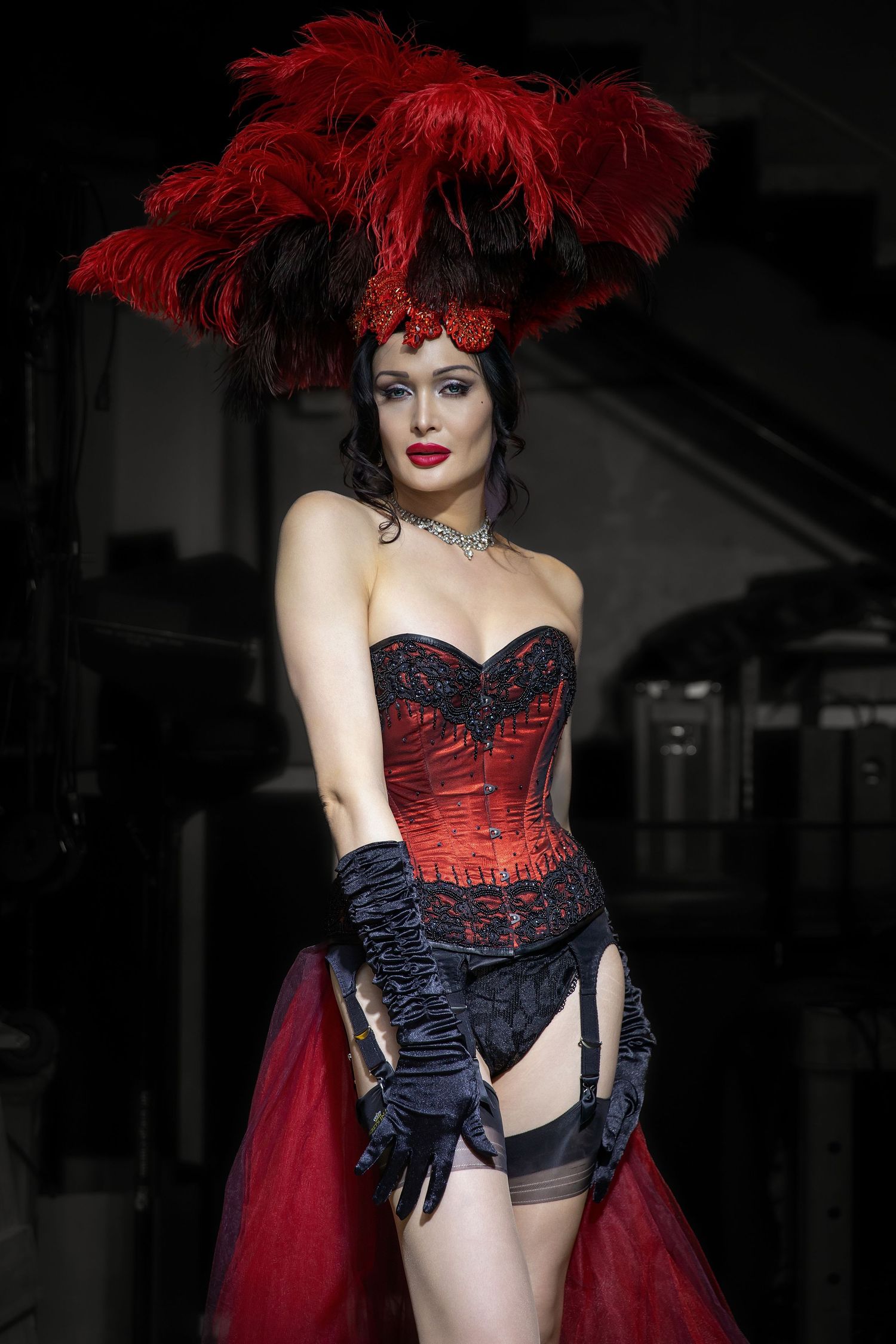 Fashion model wearing dramatic red and black corset with feathered headpiece and long gloves on dark runway.