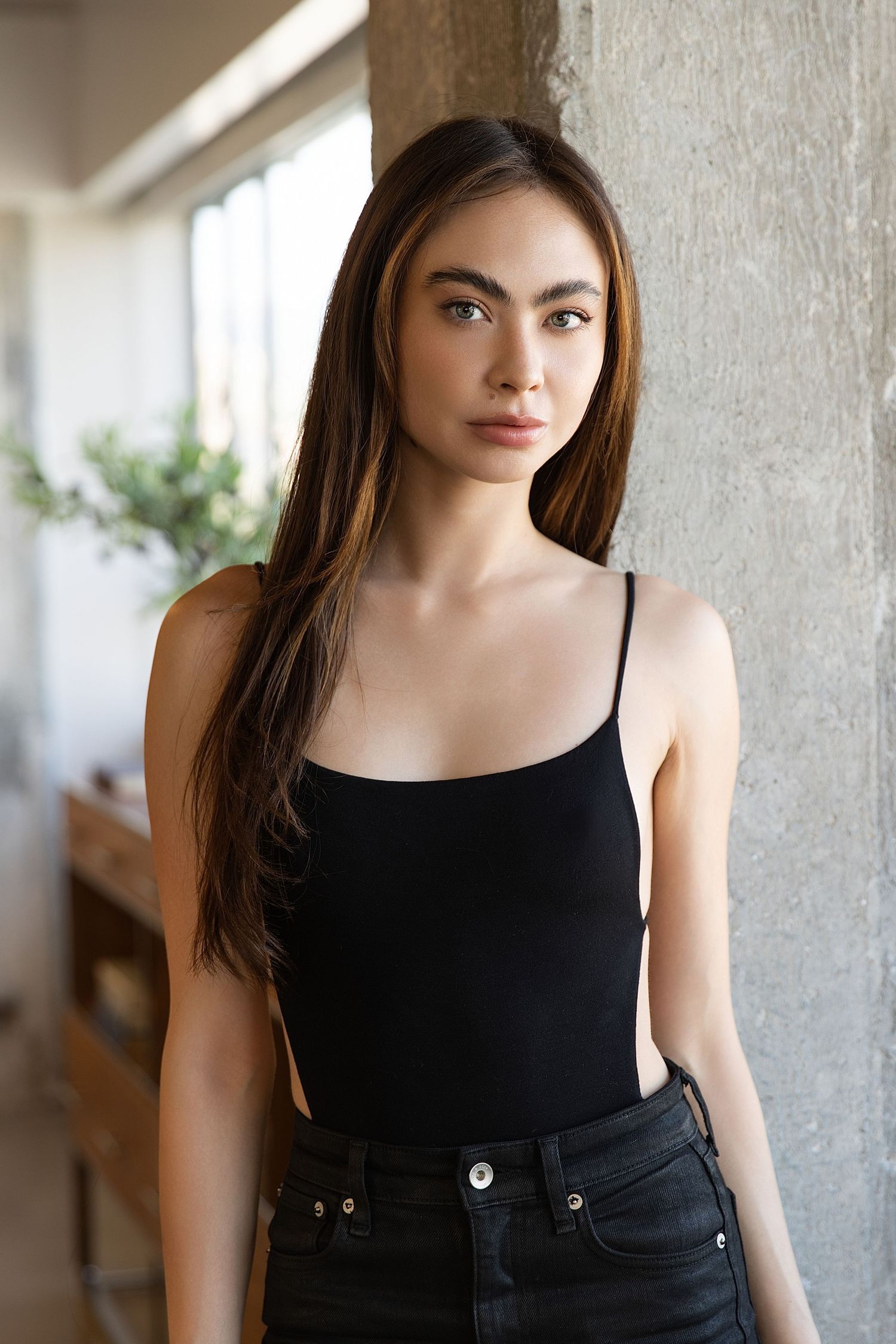 Young woman in black tank top and jeans poses against concrete wall in natural lighting.