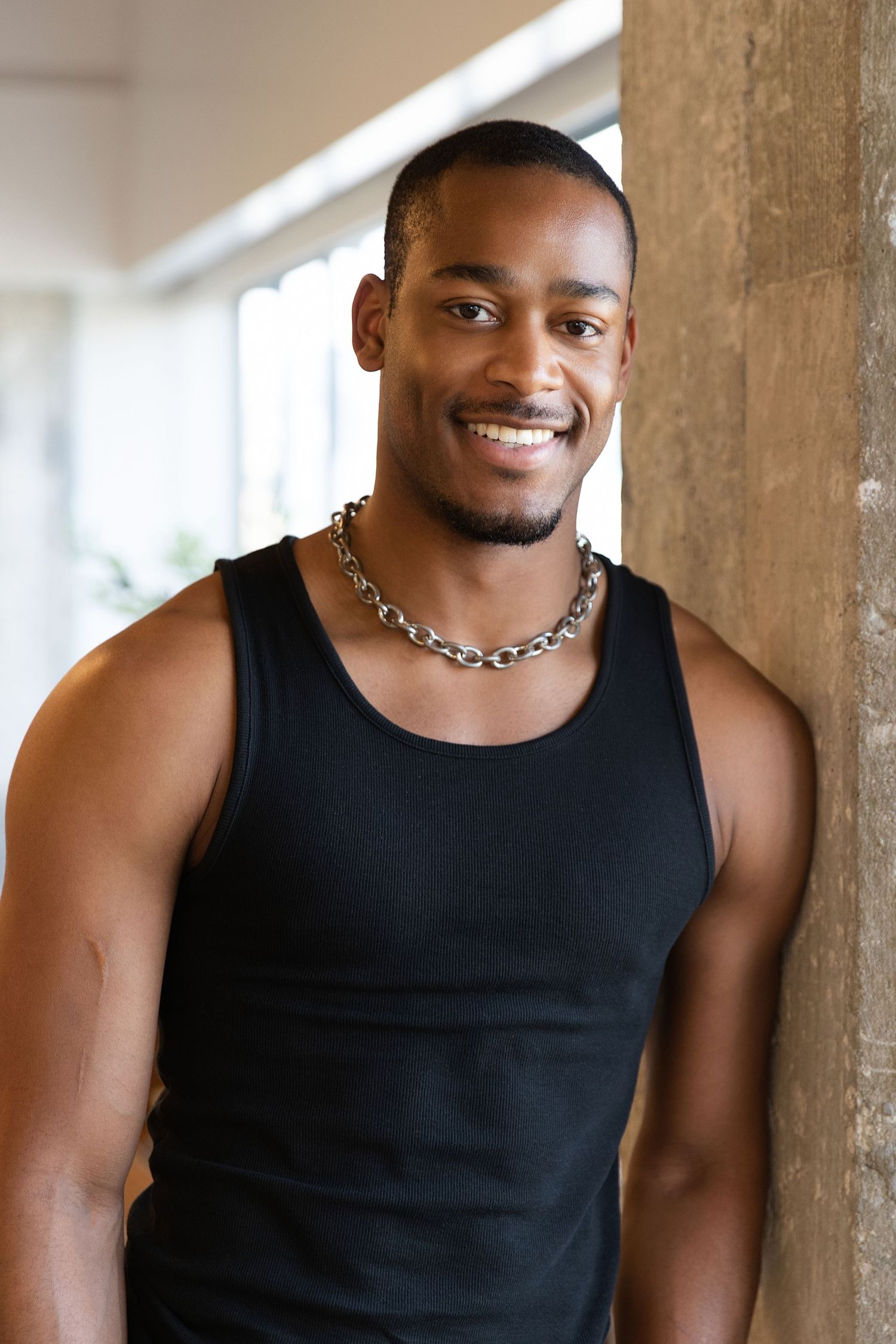 A person wearing a black tank top smiles warmly while sitting in a bright indoor setting.