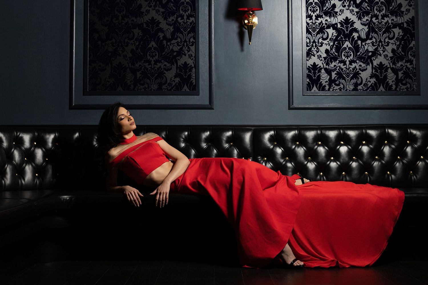 A person in a flowing red gown reclines on a black leather couch against dark damask wallpaper in moody lighting.