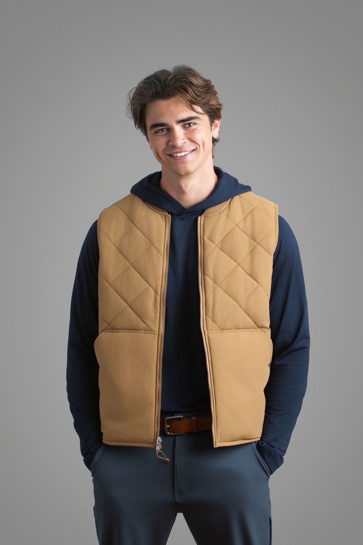 Young man smiling in a tan quilted vest over a navy hoodie with gray pants on a gray background.