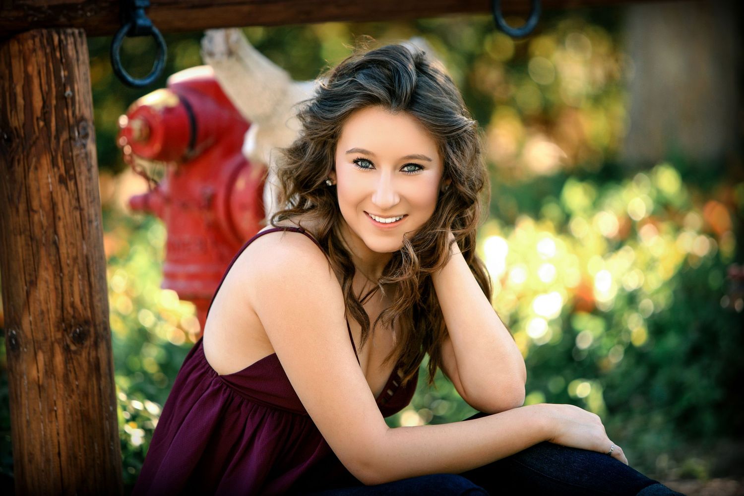 Young woman in burgundy dress smiling outdoors near a red fire hydrant in a lush green setting.