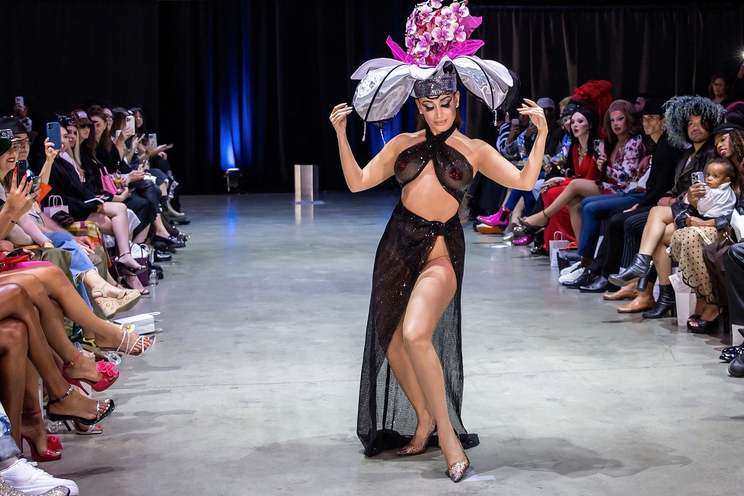 Models walk the runway in black evening wear during a fashion show with dramatic blue lighting and audience seating.
